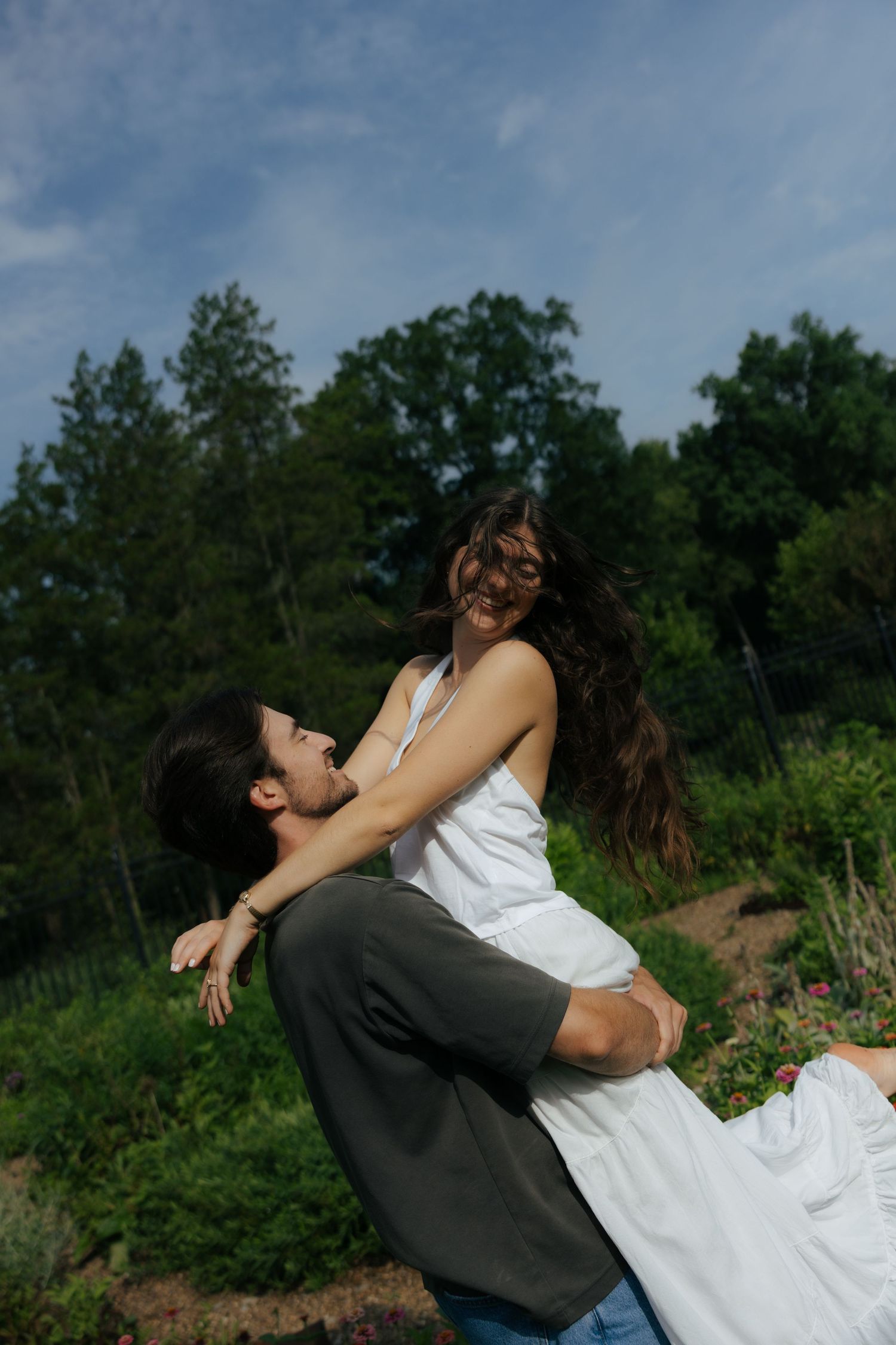 Man joyfully lifting woman in white dress outdoors with trees and blue sky behind them.