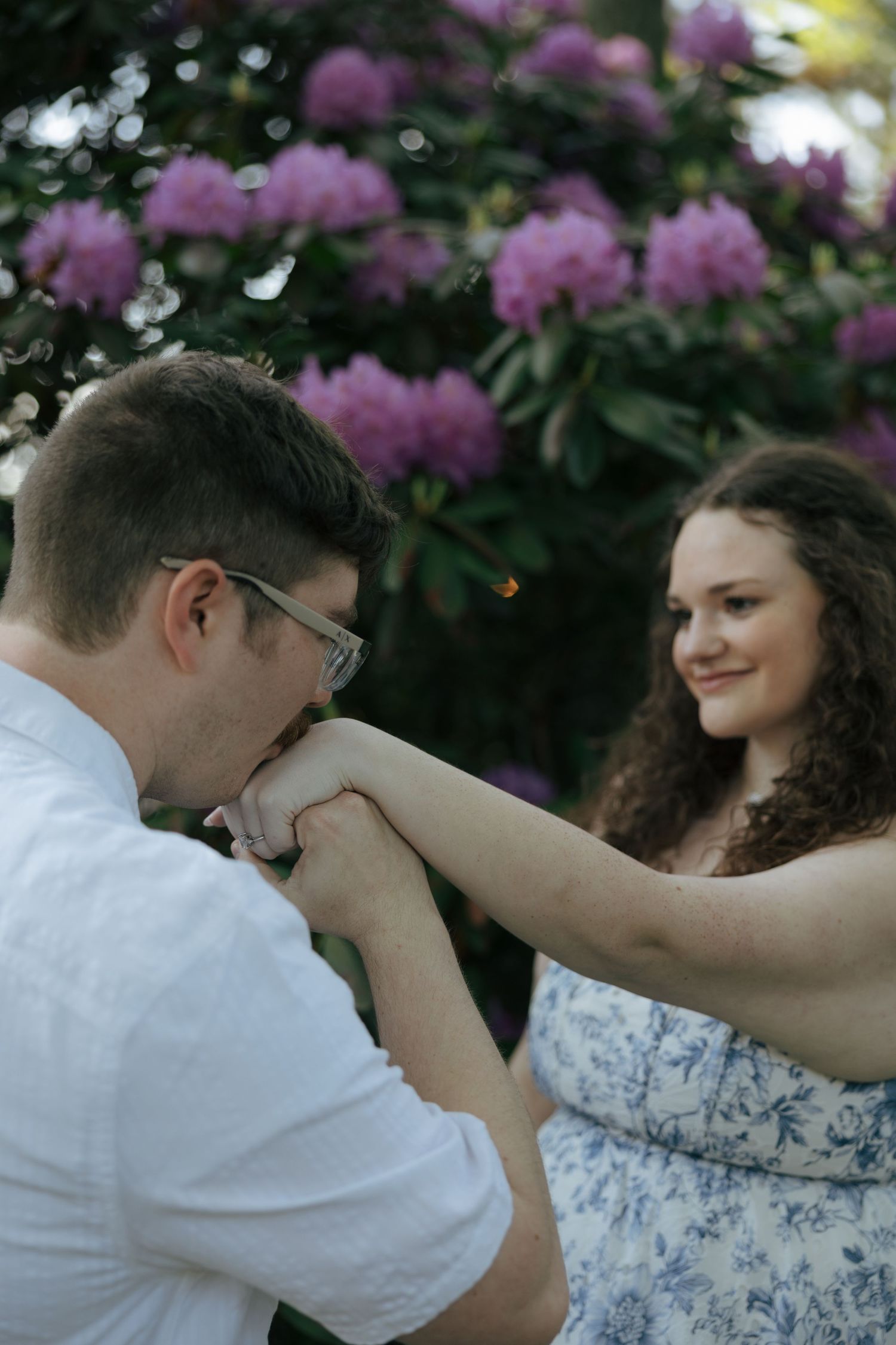 Man kissing woman's hand in front of blooming purple rhododendron flowers.