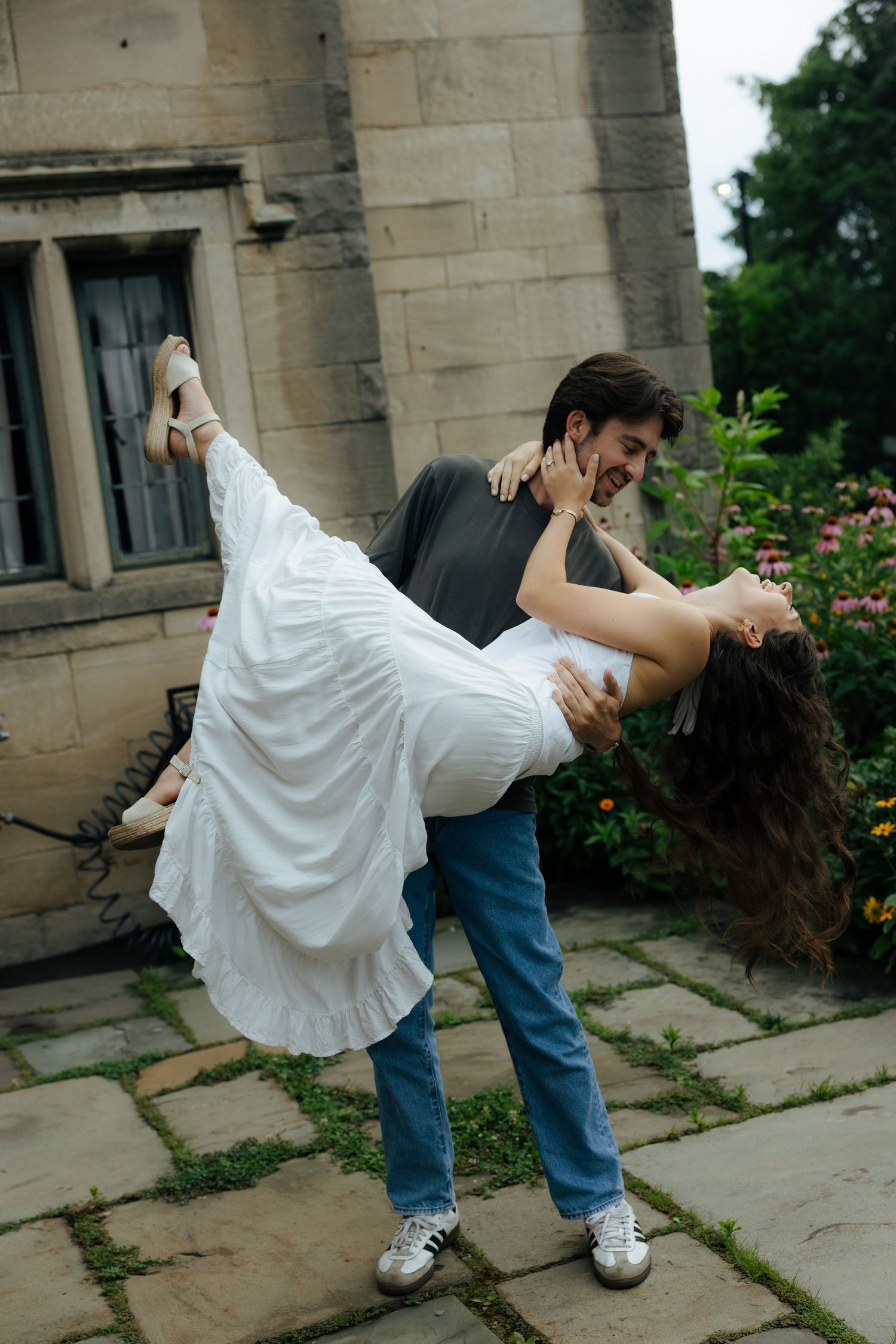 A man dips a woman in a white dress while dancing outdoors near a stone building surrounded by greenery.