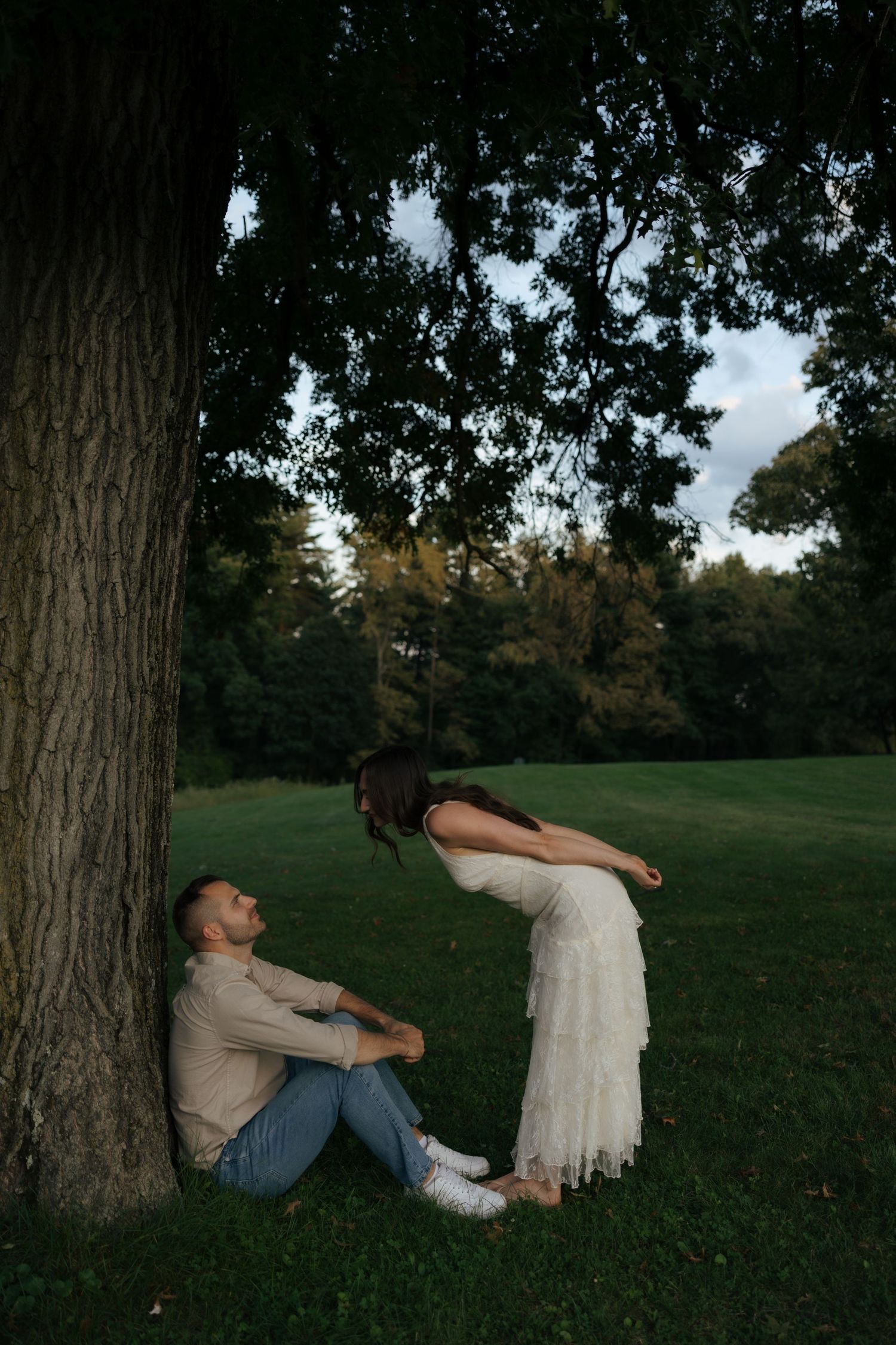 Groom sits against large tree as bride in white lace dress leans over him playfully.
