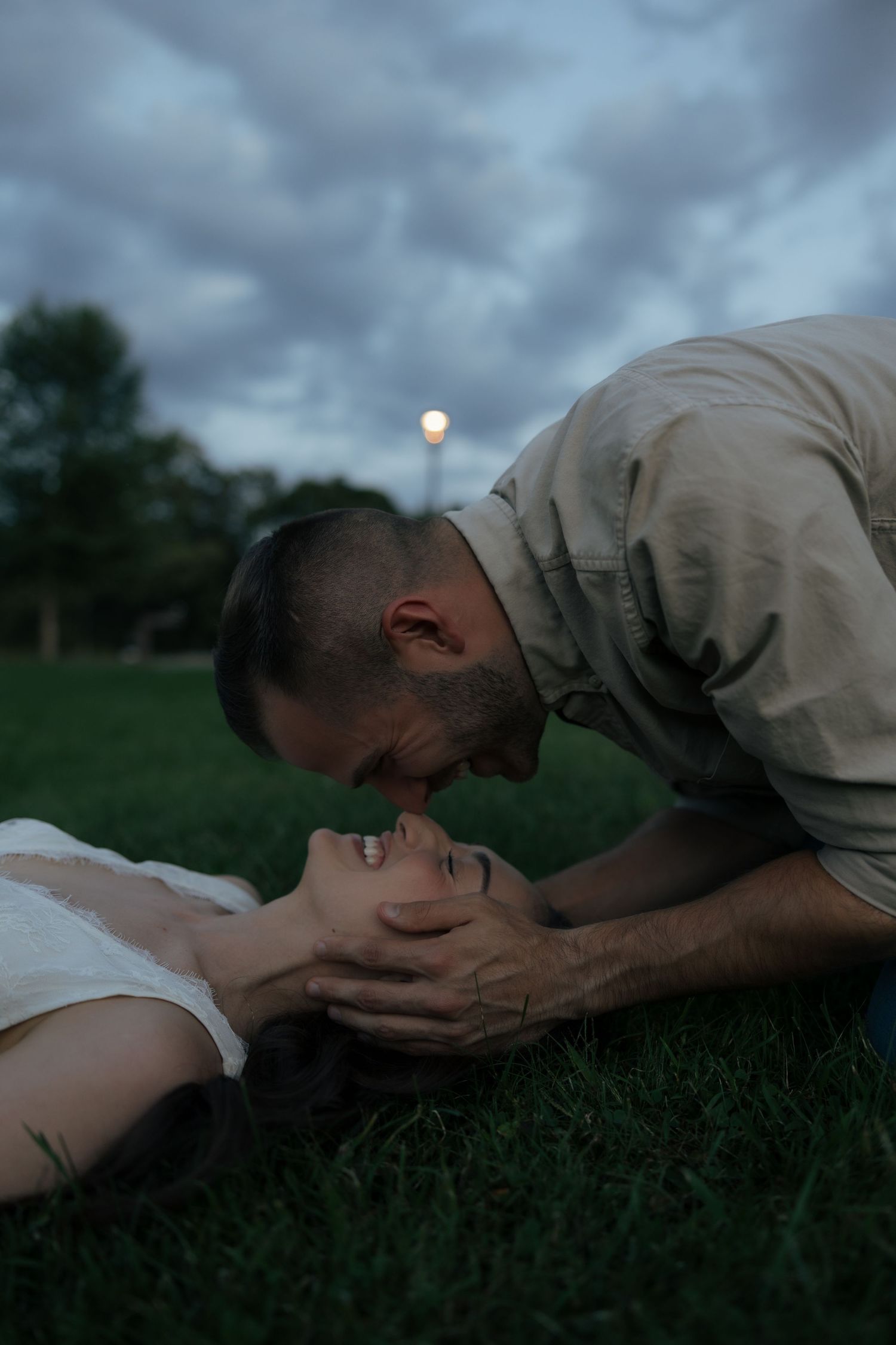 Romantic couple at dusk, man leaning down to kiss woman lying on grass under cloudy sky.
