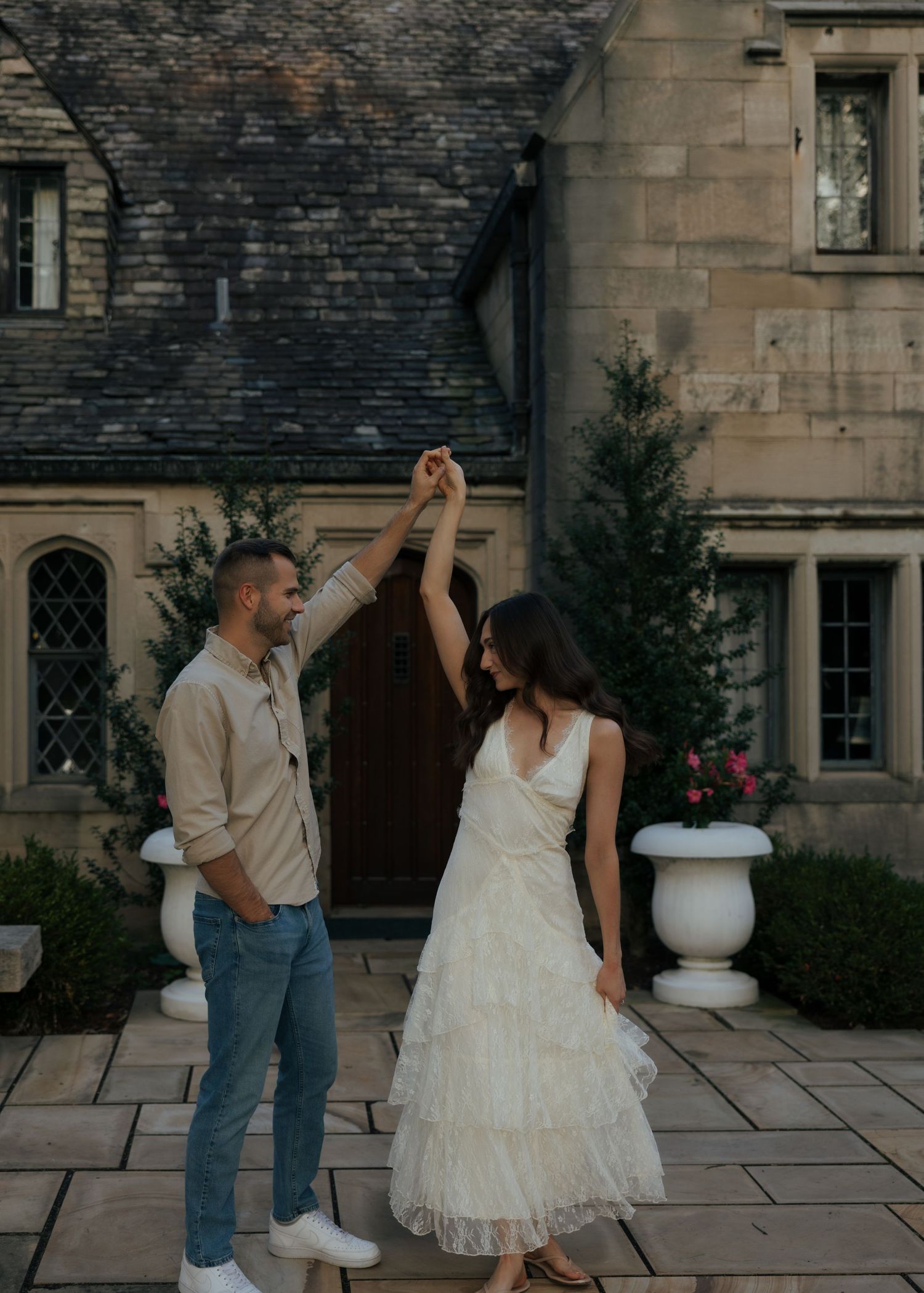 Couple dancing together outside a stone manor, woman in white tiered dress being twirled by man in beige shirt and jeans.
