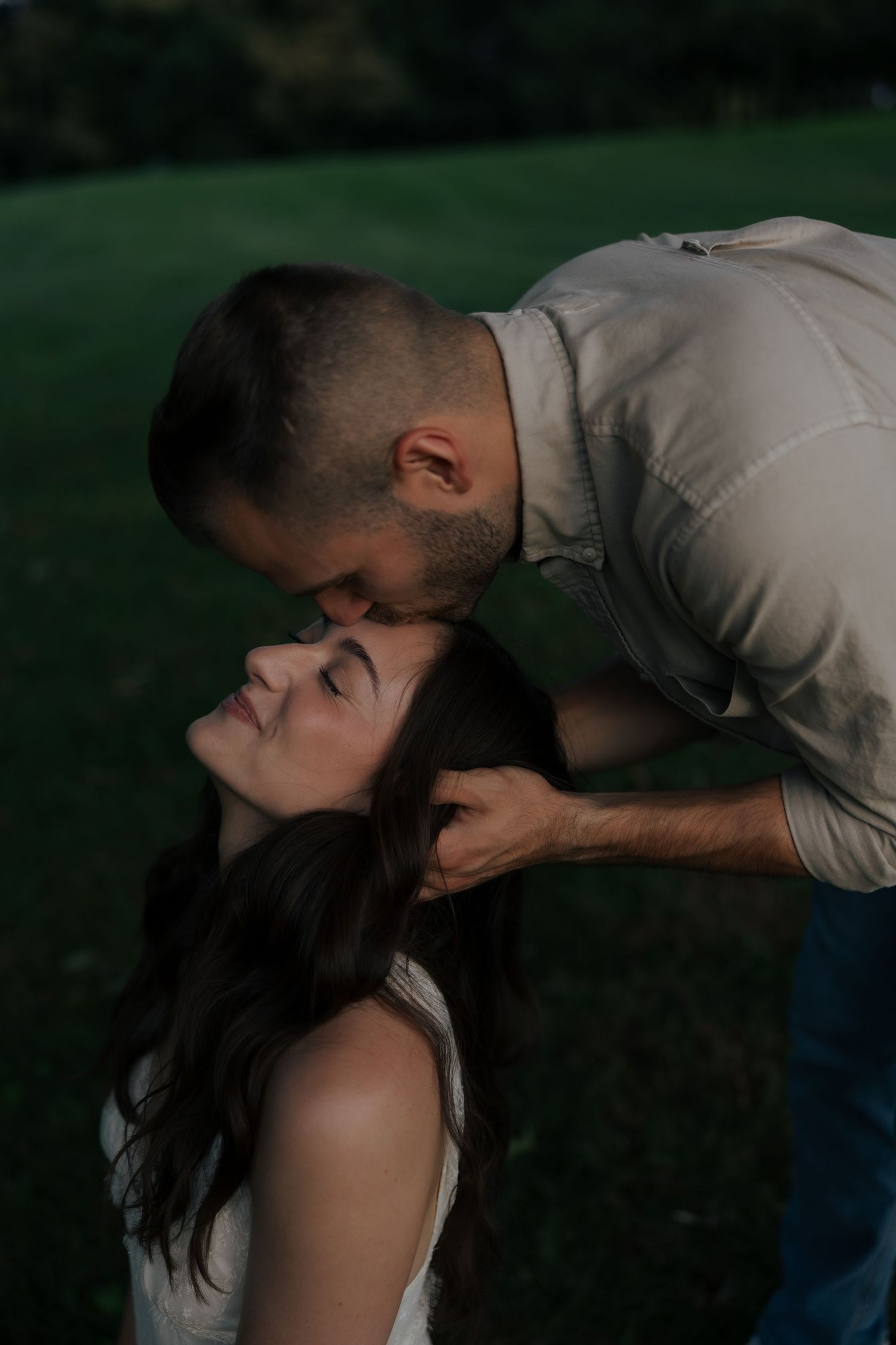A man tenderly kisses a woman on the forehead while holding her hair back in a romantic outdoor setting.