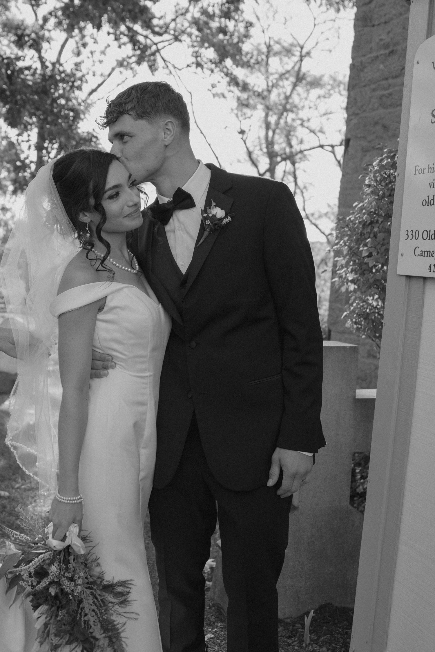 Groom kisses bride on forehead outside church sign.