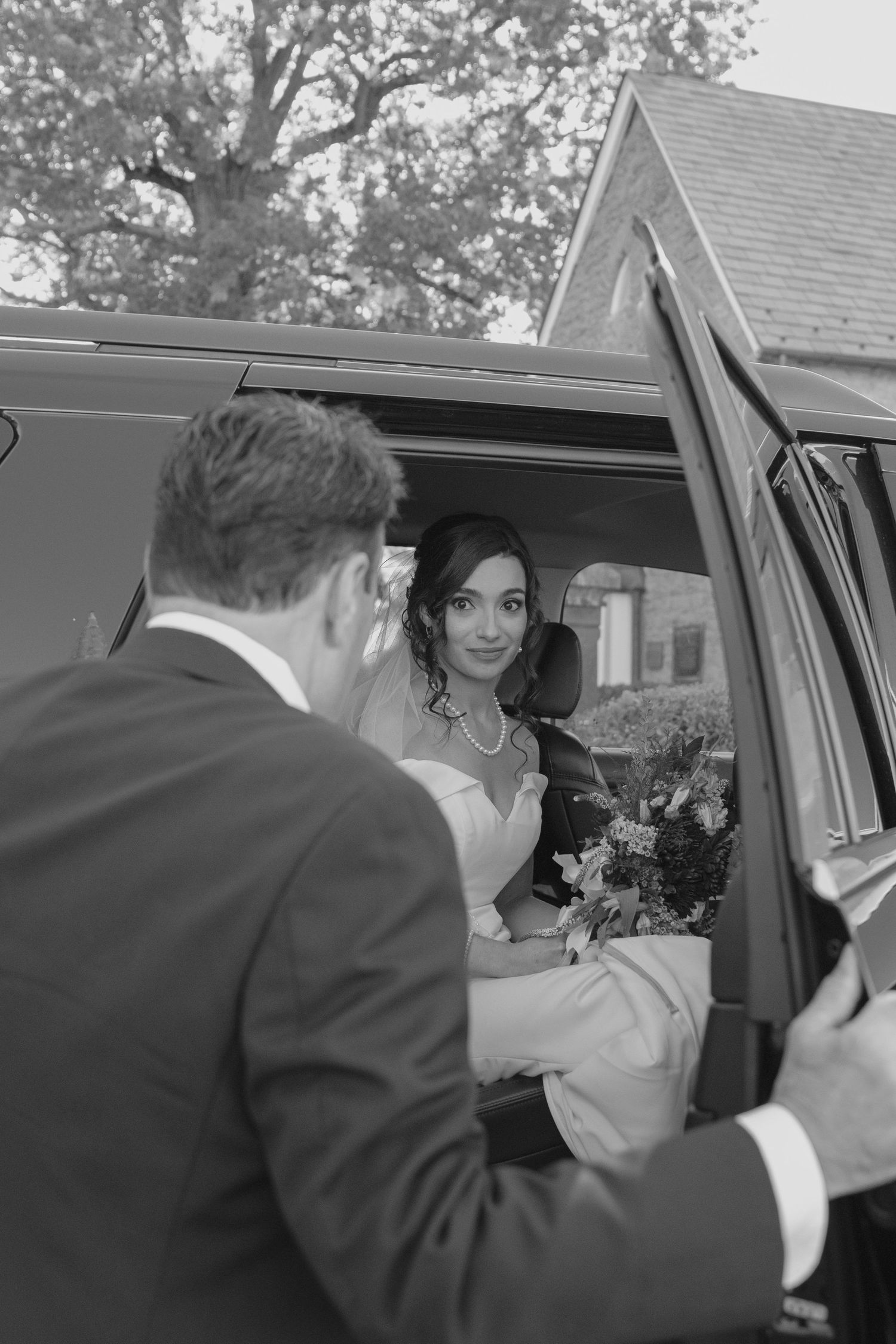 Bride sitting in wedding car holding bouquet, smiling at groom in black suit outside vehicle, black and white photo.