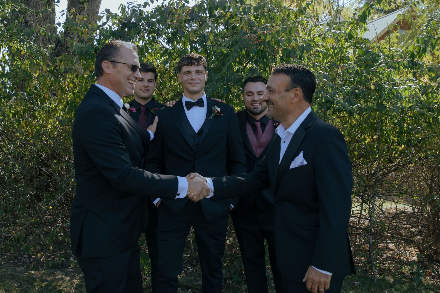 Two men in tuxedos shake hands while groomsmen smile in background at wedding.