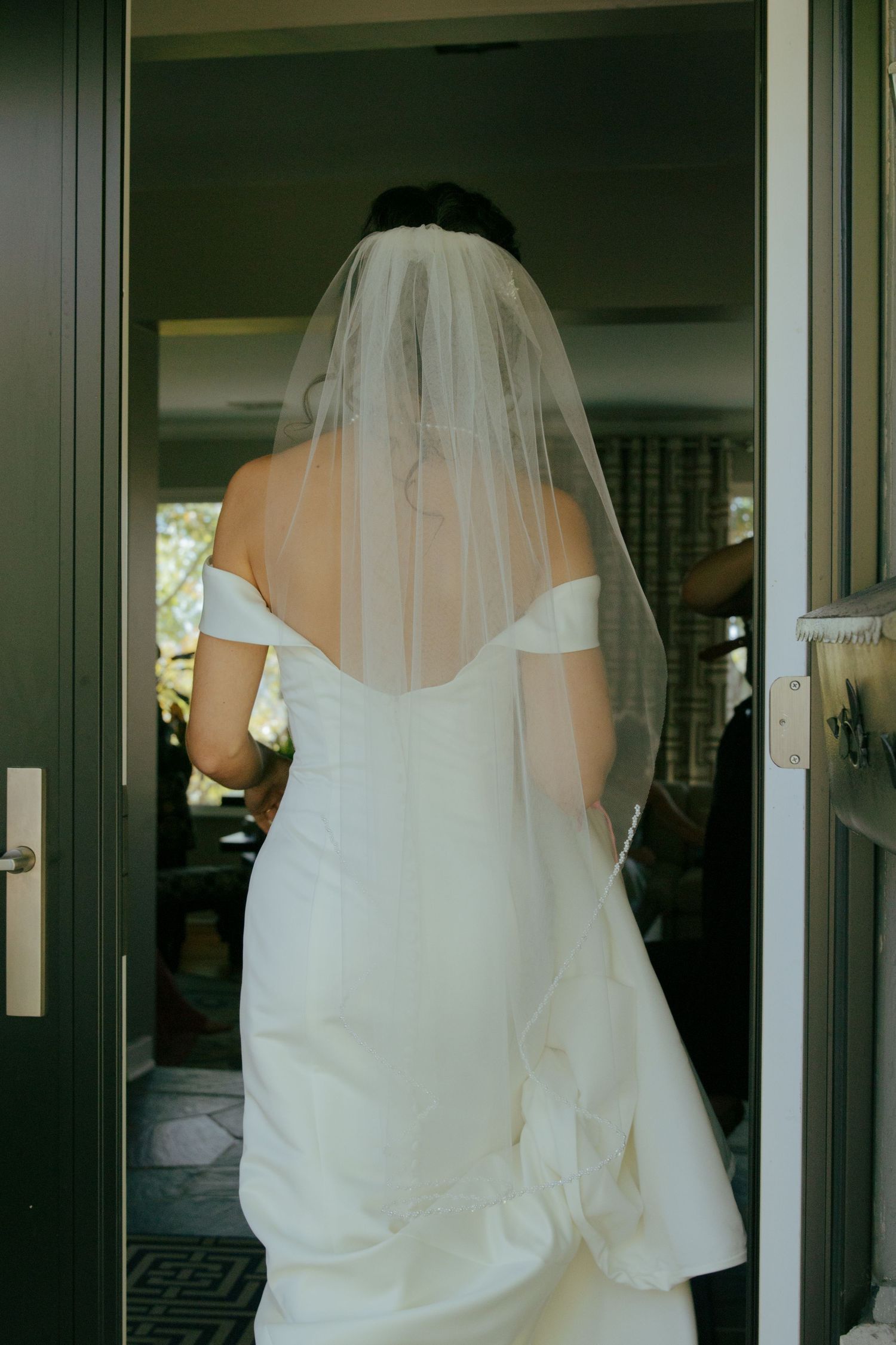 Bride in white off-shoulder gown and veil seen from behind standing in doorway.