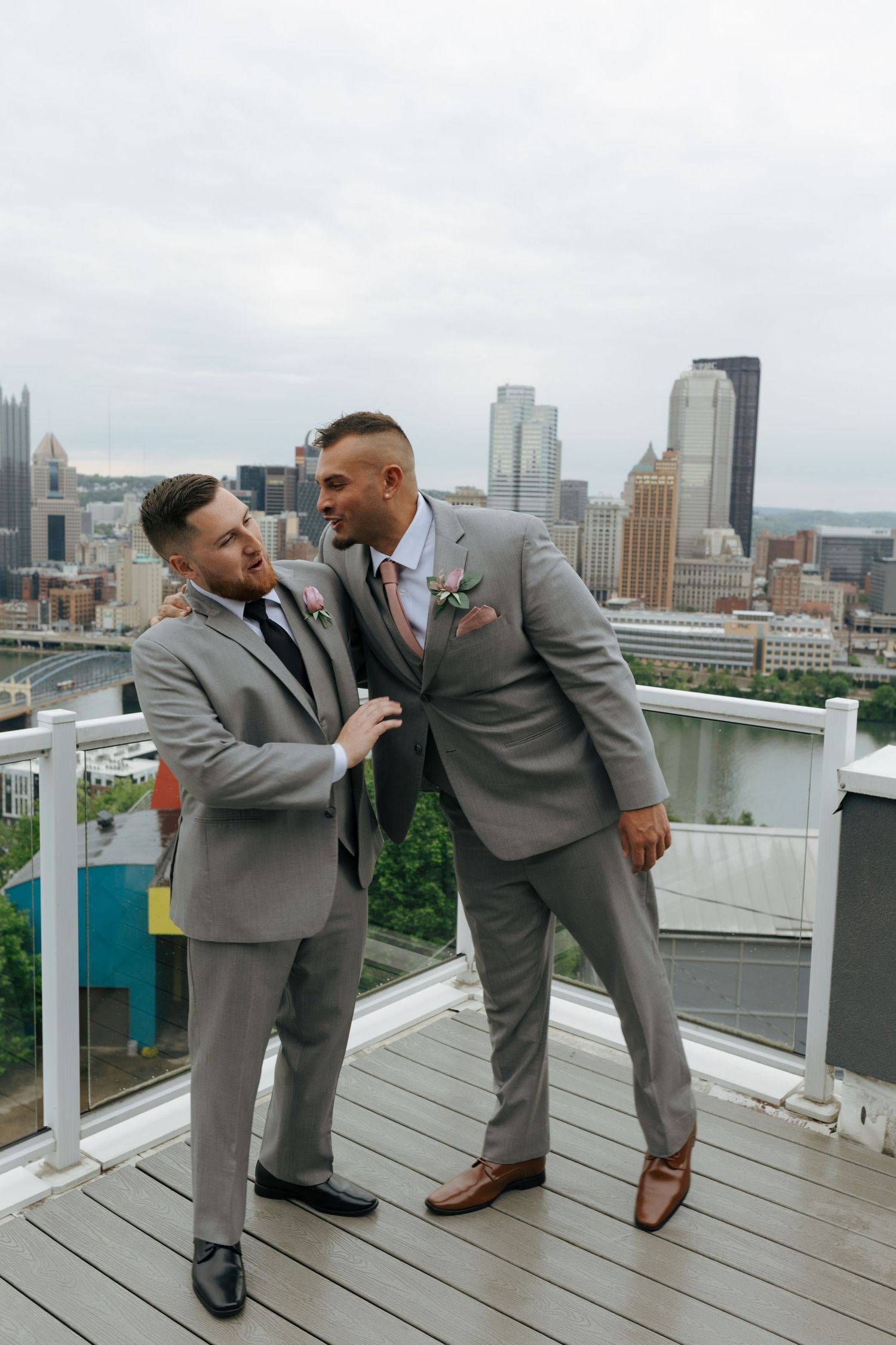 Two groomsmen in grey suits share a laugh on a rooftop overlooking Pittsburgh skyline.