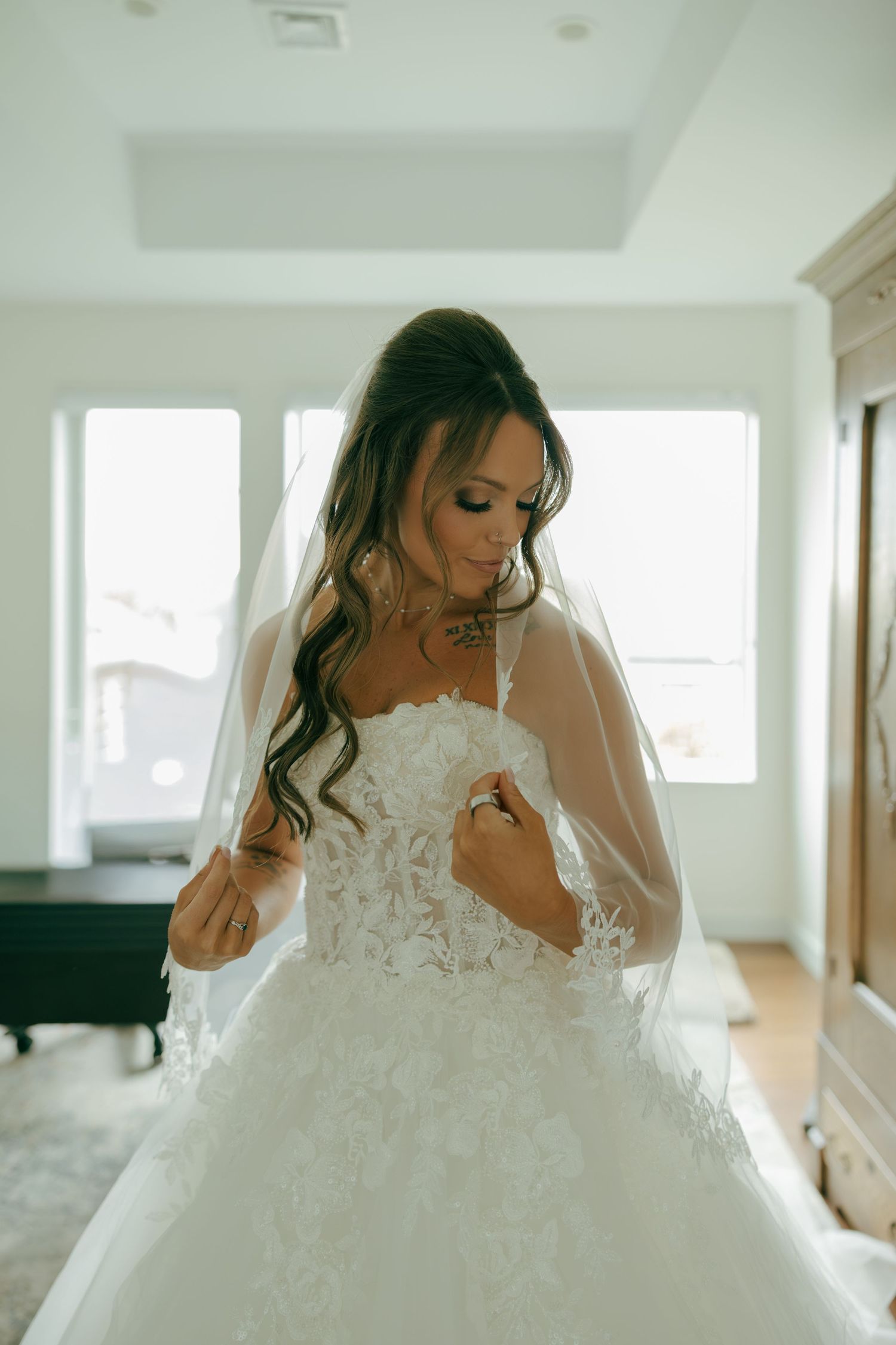 Beautiful bride in strapless lace ball gown with veil looking down in a bright sunlit bedroom.