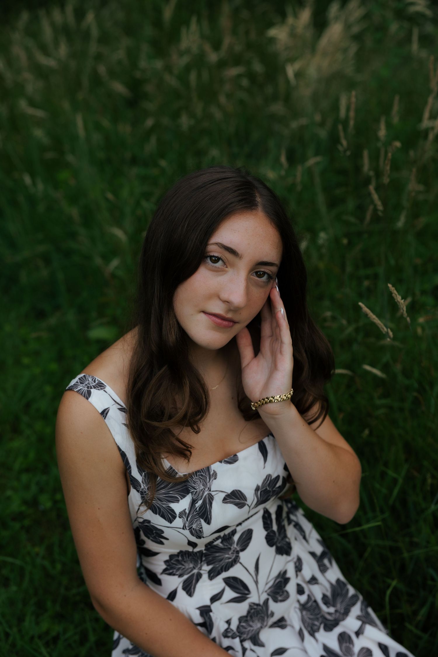 Young woman in floral dress sitting in tall grass, hand resting on cheek, serene outdoor portrait.