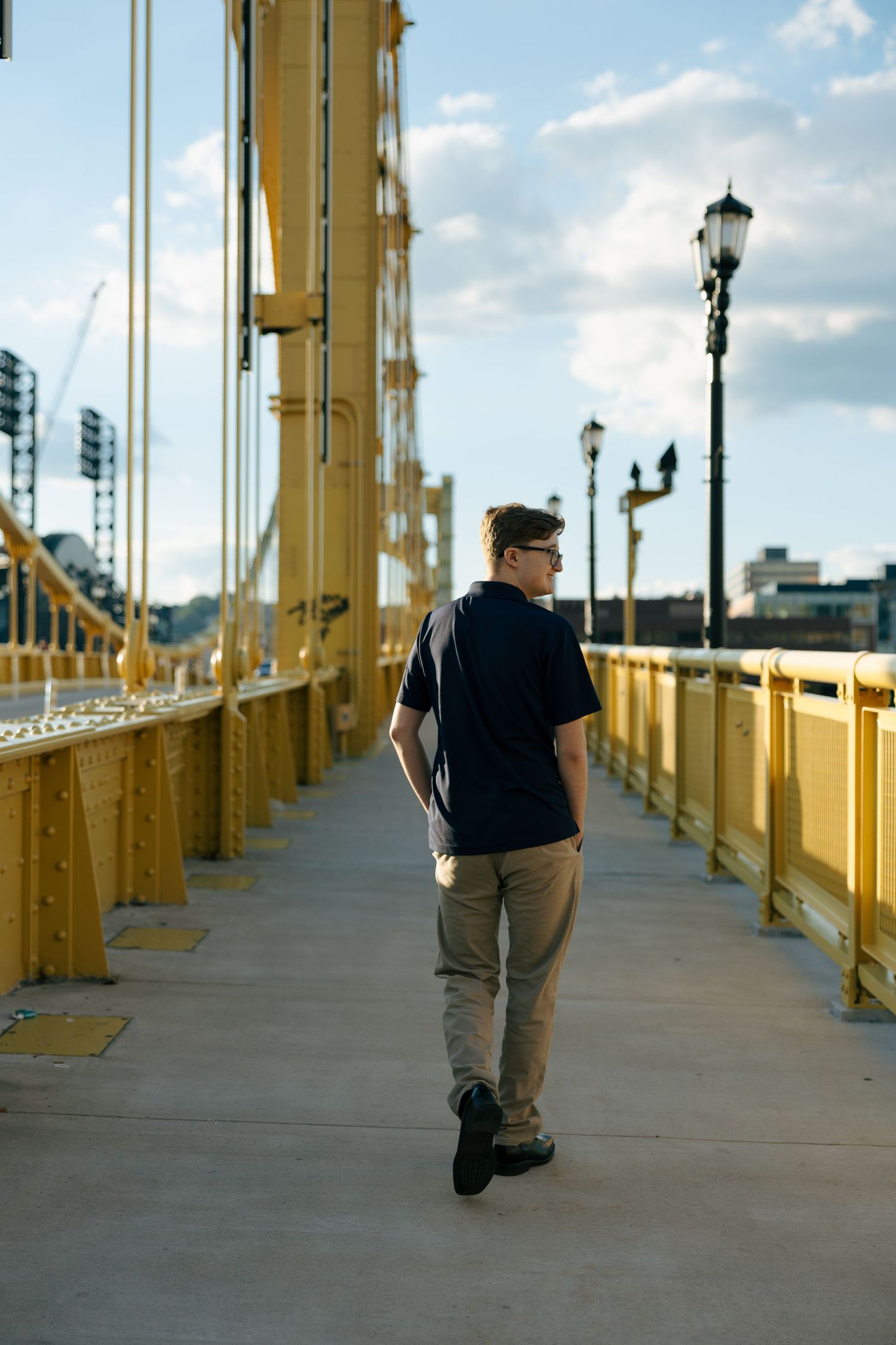 Young man in navy polo and khakis walking across a yellow bridge in Pittsburgh on a sunny day.