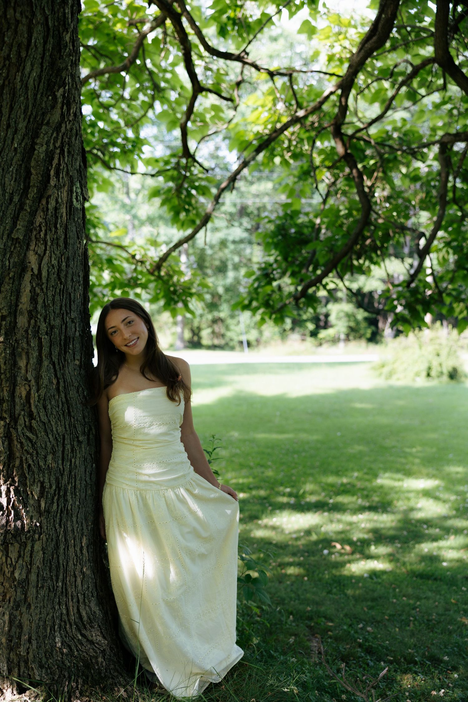 Young woman in elegant white gown leaning against large tree in lush green park setting.