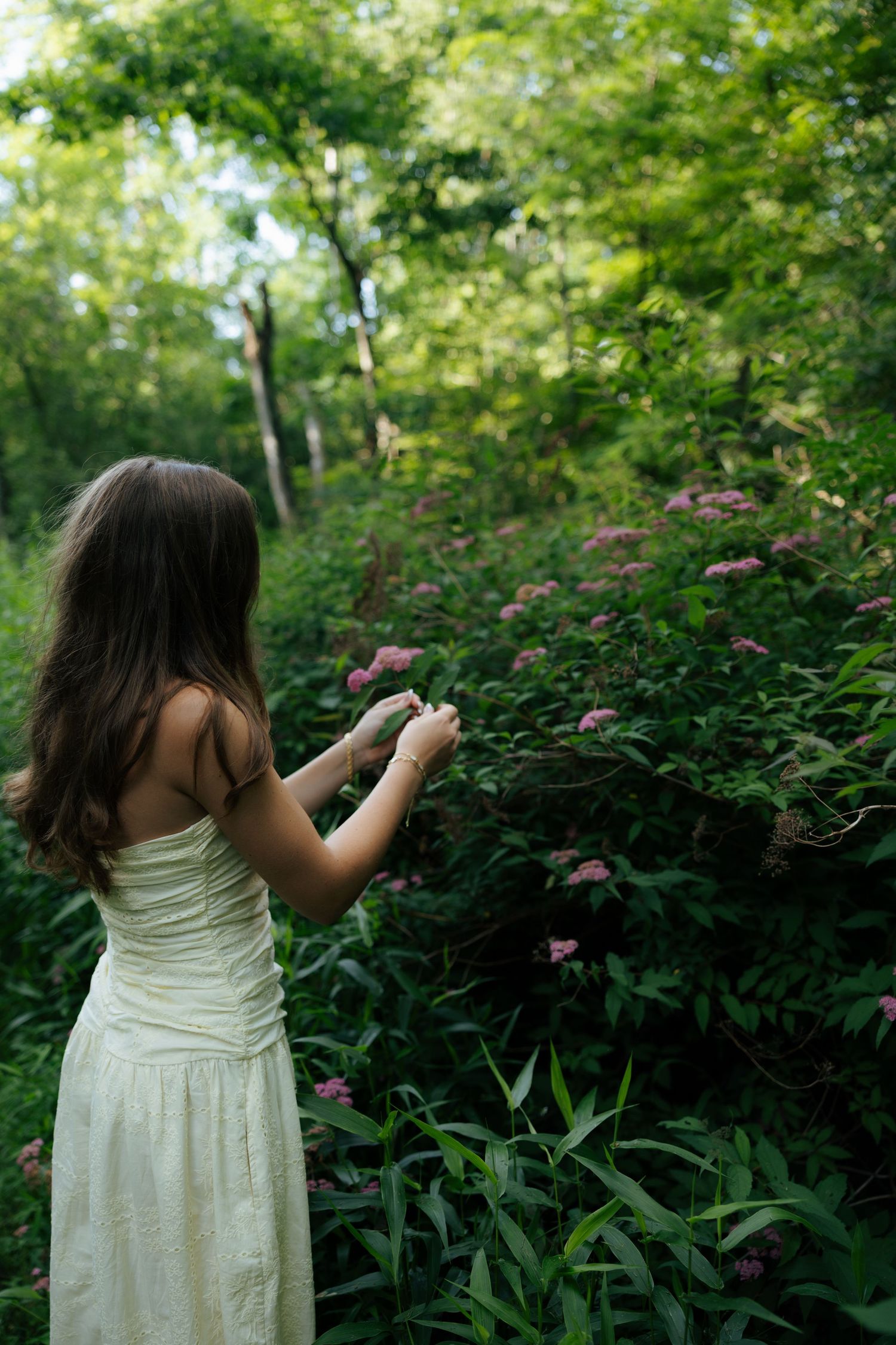 Woman in white dress reaching toward pink wildflowers in forest.