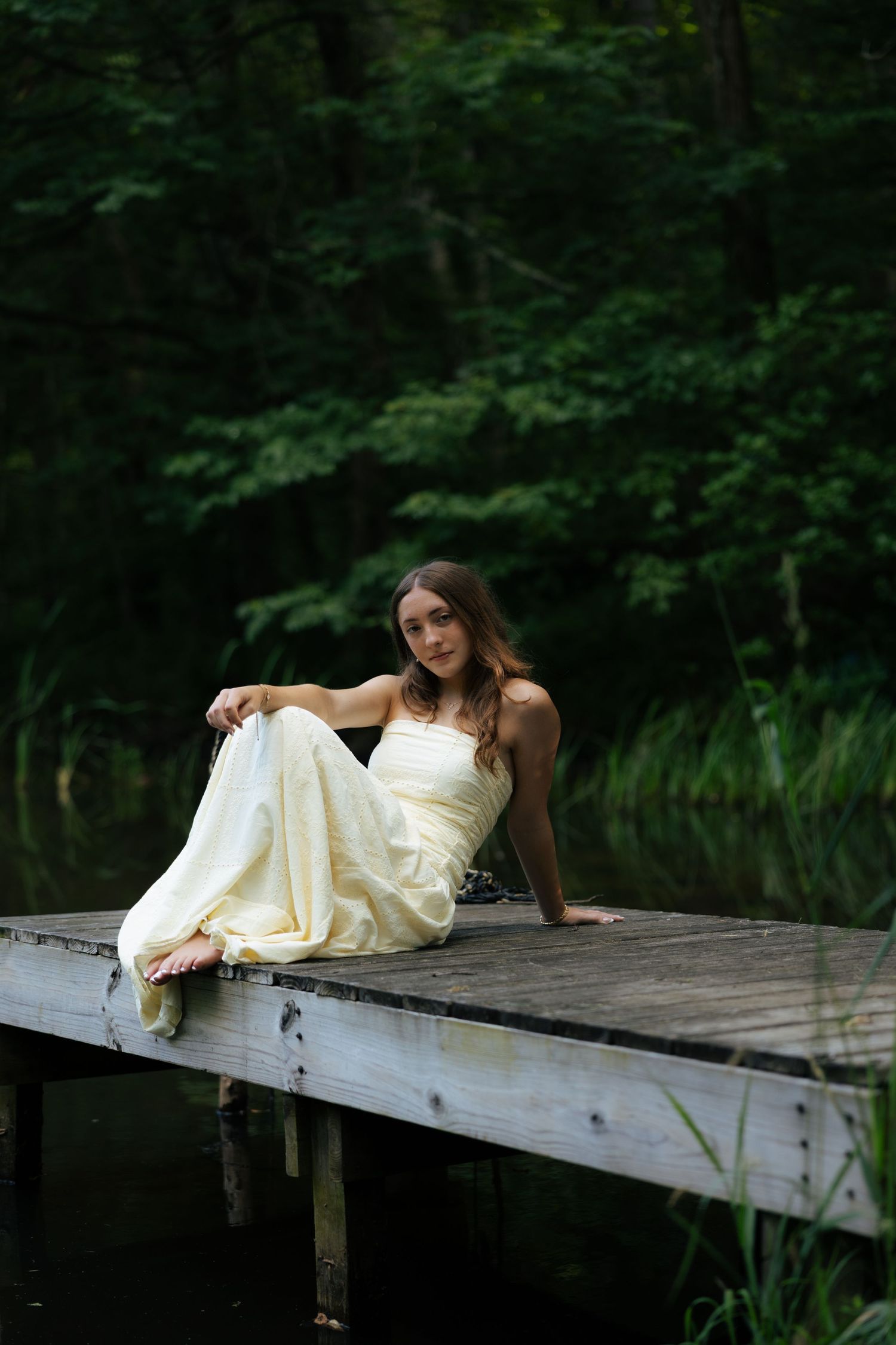 Young woman in cream strapless gown sitting gracefully on wooden dock by dark forest lake.