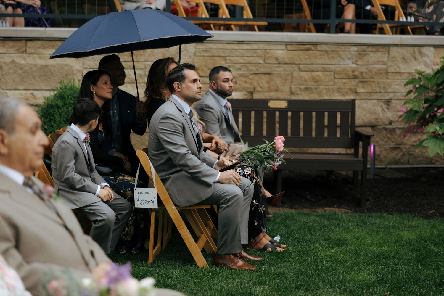 Wedding guests in gray suits sit on wooden chairs at outdoor ceremony, one holding umbrella.