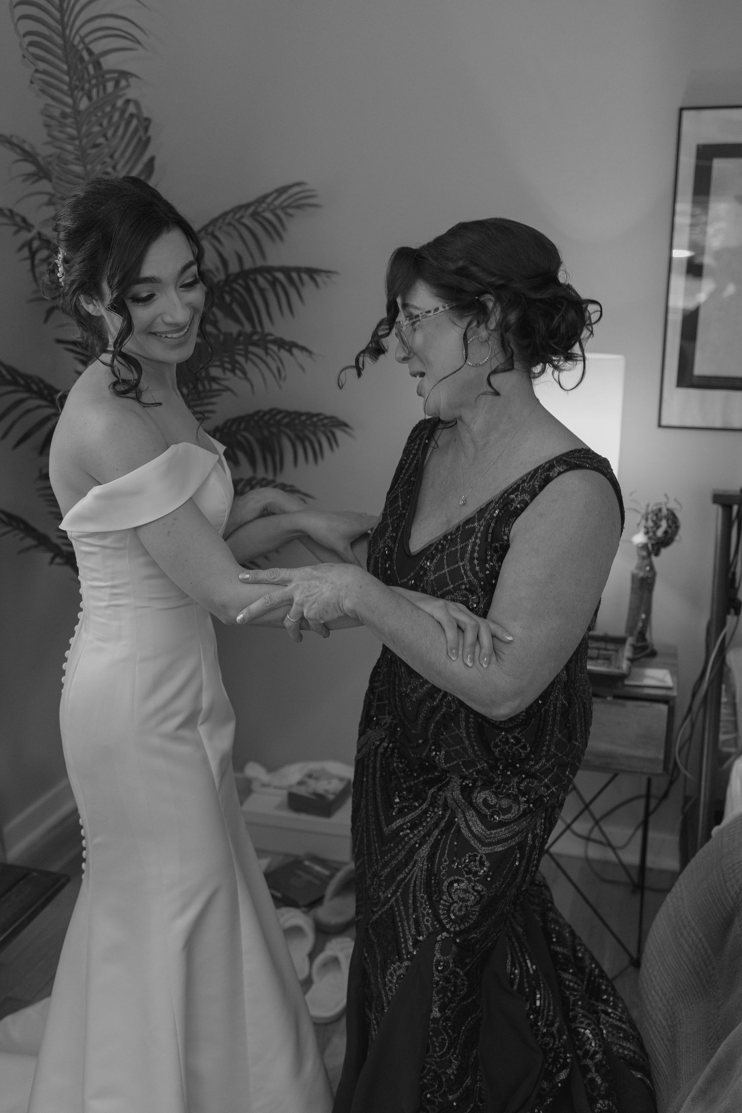 Black and white photo of mother helping bride into her off-shoulder white wedding gown.
