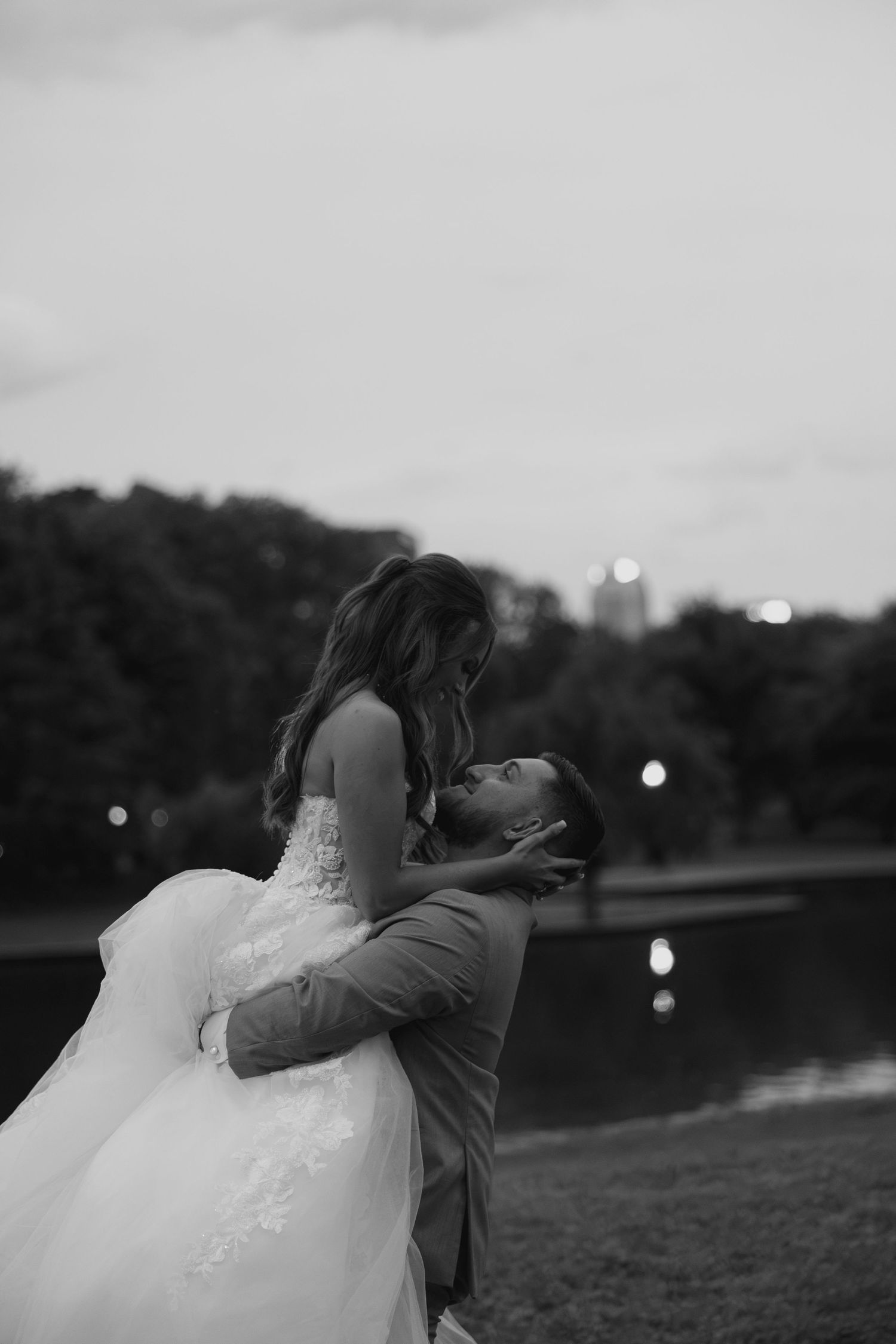 Black and white photo of groom lifting bride in wedding gown near a reflective pond at dusk.