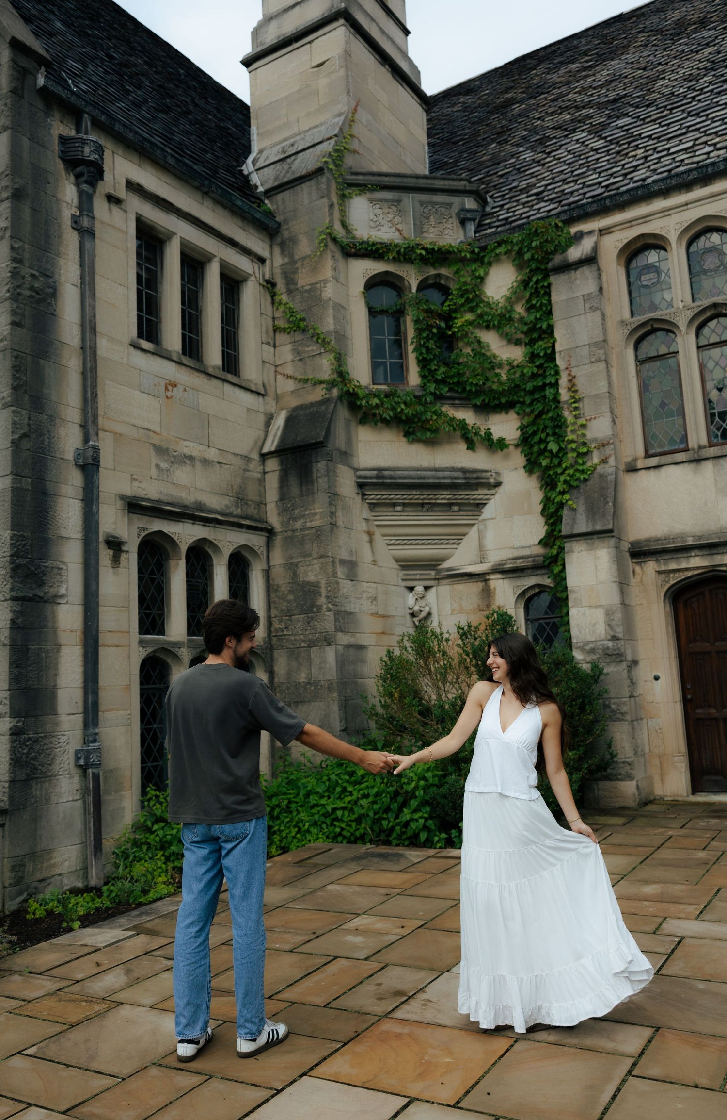 Couple holding hands in courtyard of ivy-covered gothic stone building, woman in flowing white dress.