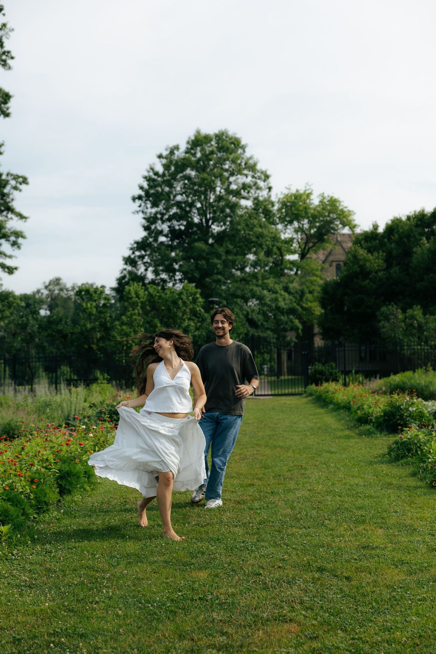 A woman in a flowing white dress runs barefoot through a lush green garden path as a man follows behind her.