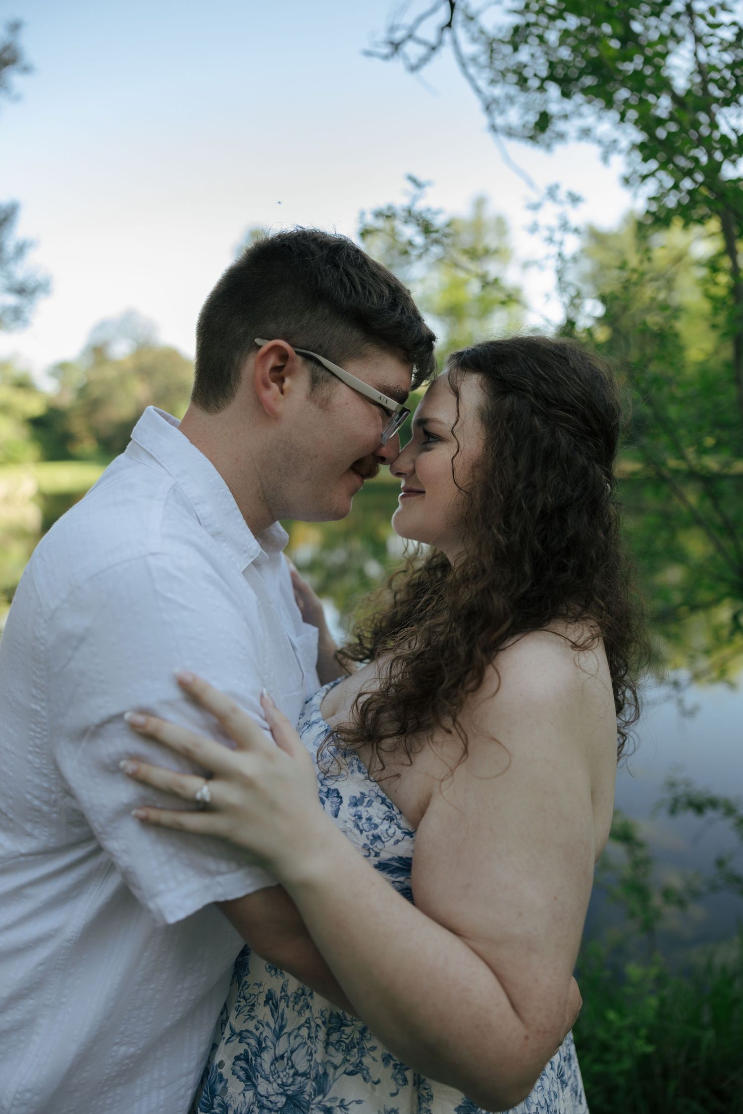Couple sharing a tender moment touching foreheads outdoors near a pond surrounded by lush green trees.