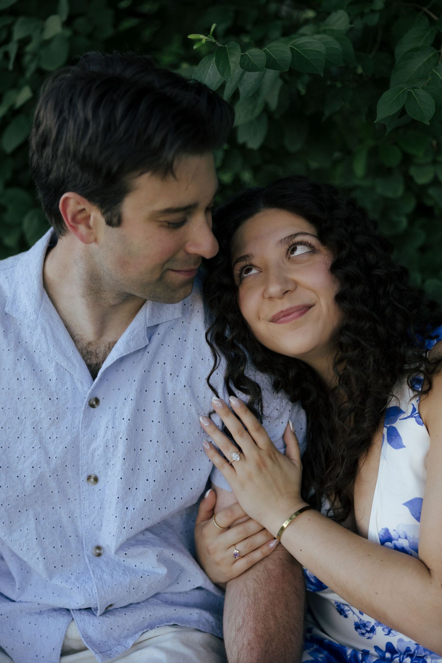 Couple sitting together outdoors, woman showing engagement ring while smiling up at man near green foliage.