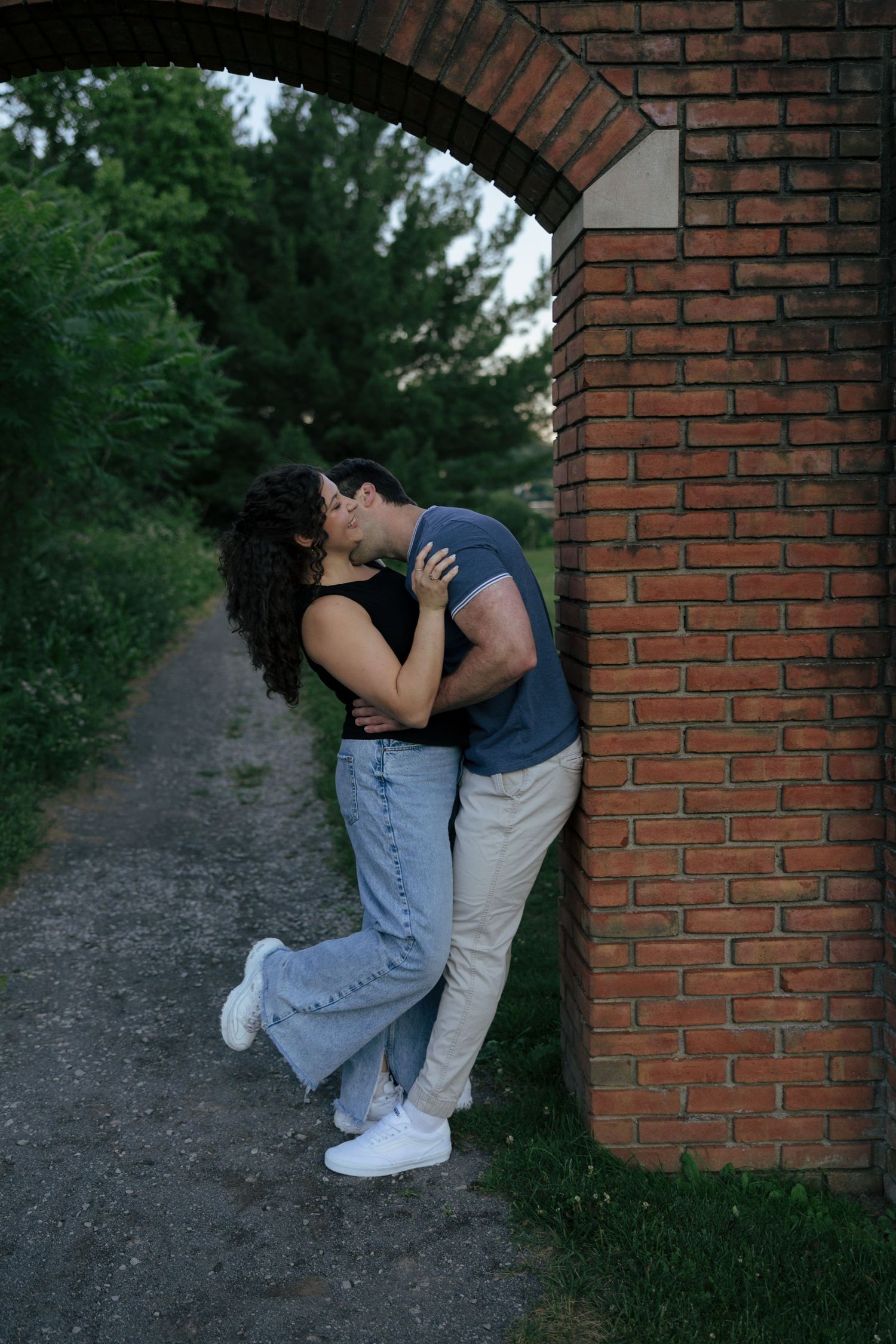 Couple sharing a romantic kiss against a brick arch surrounded by lush greenery.