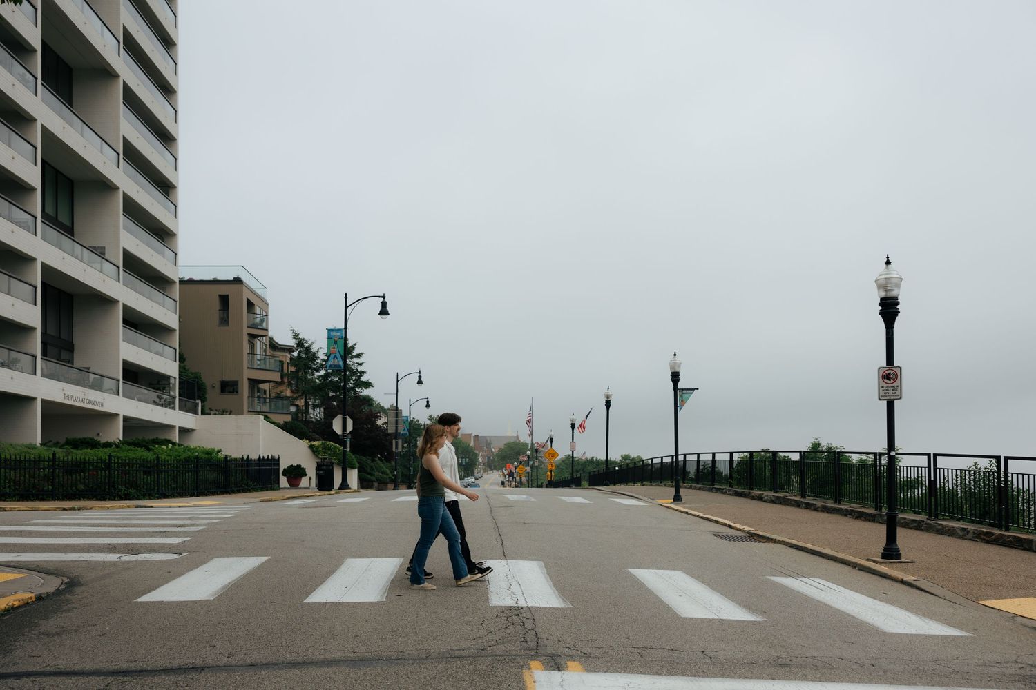 A man crosses a zebra crosswalk on a quiet urban street beside a tall building on an overcast day.