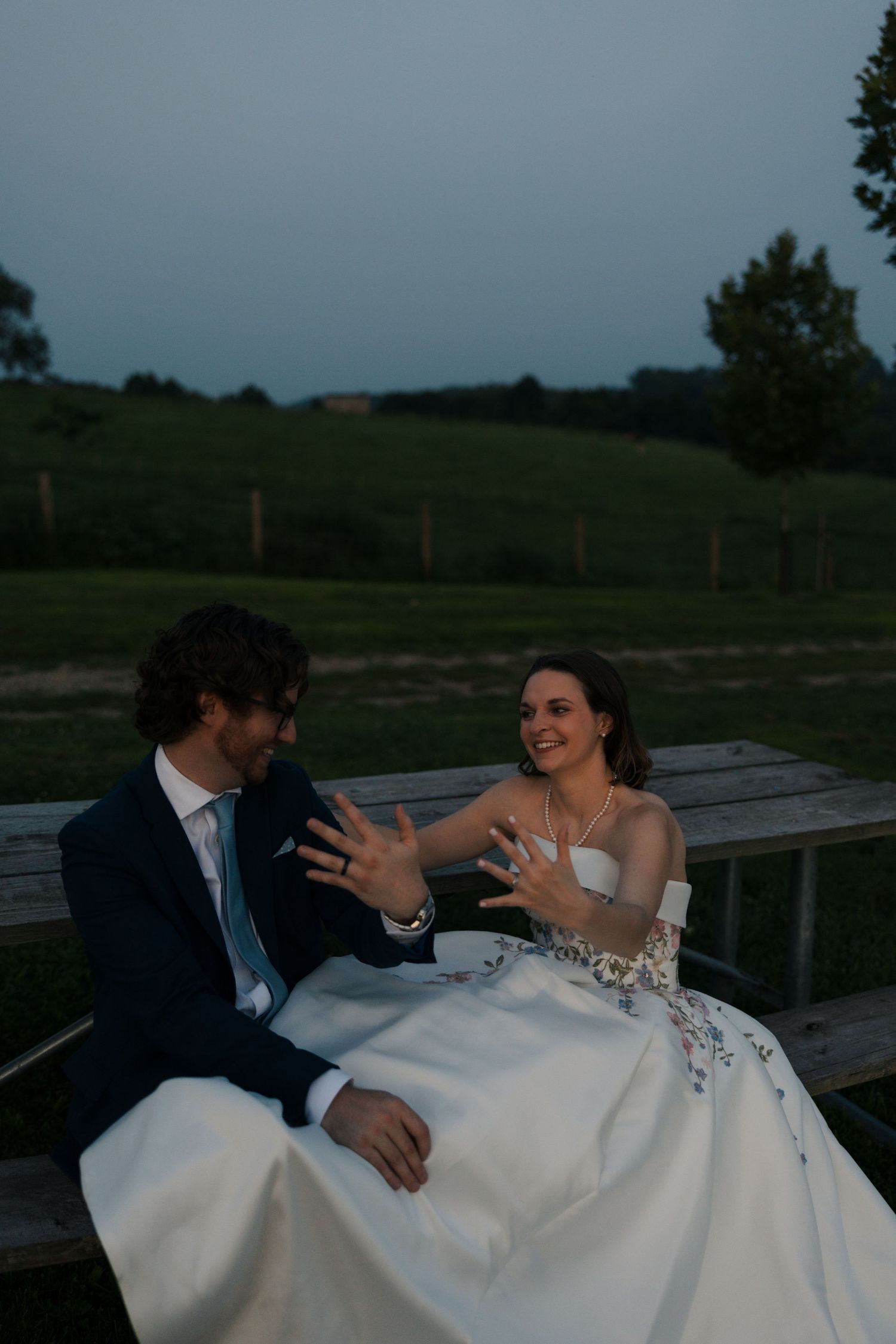 Newlyweds show off wedding rings while seated on a bench at dusk in a scenic green field.