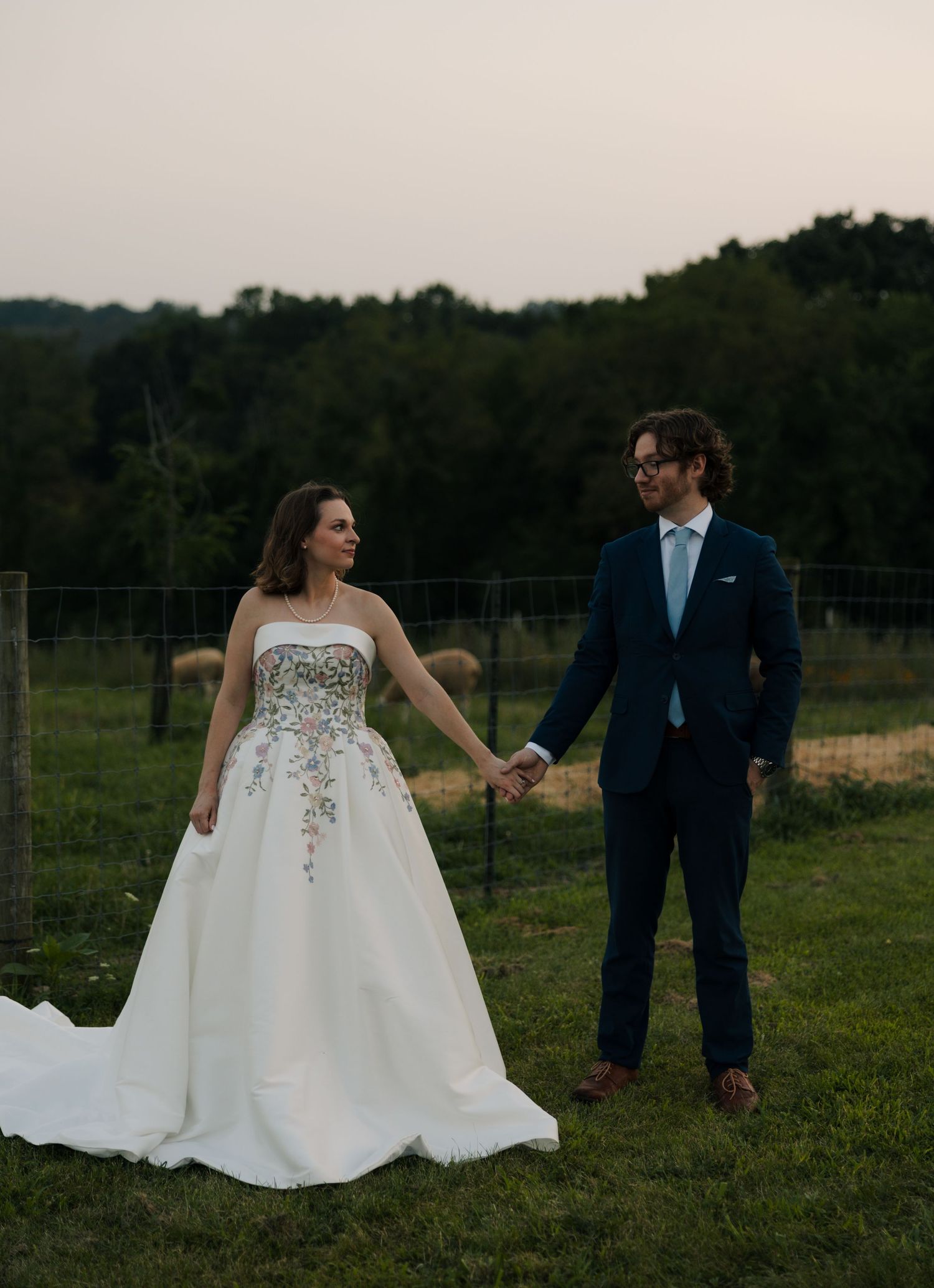 Bride and groom hold hands near farm fence at dusk, bride wearing strapless floral ballgown wedding dress.