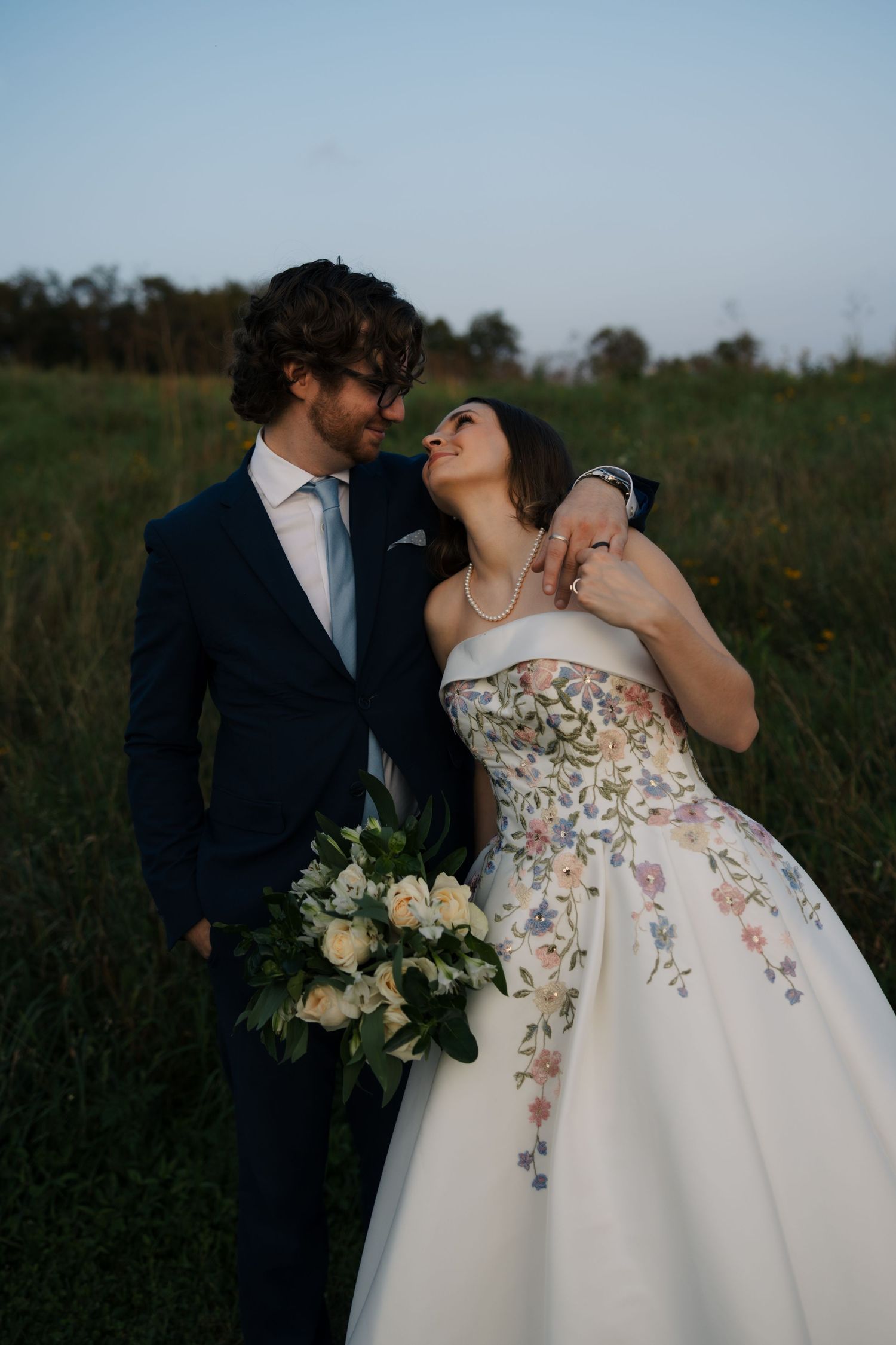 Bride and groom share a romantic moment in a field, she holds white bouquet in floral embroidered gown.