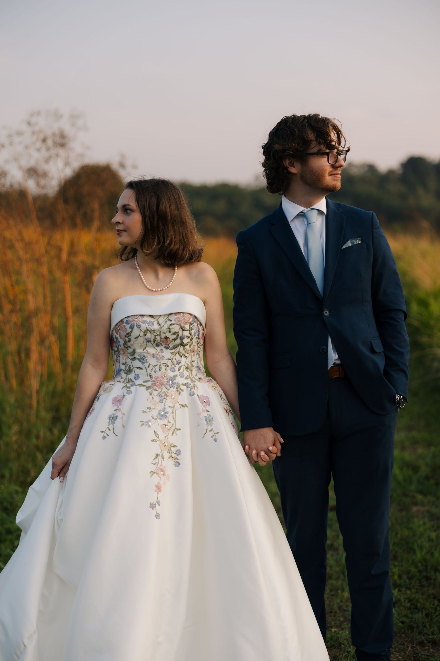Bride in floral embroidered wedding gown holds hands with groom in navy suit in golden field at sunset.