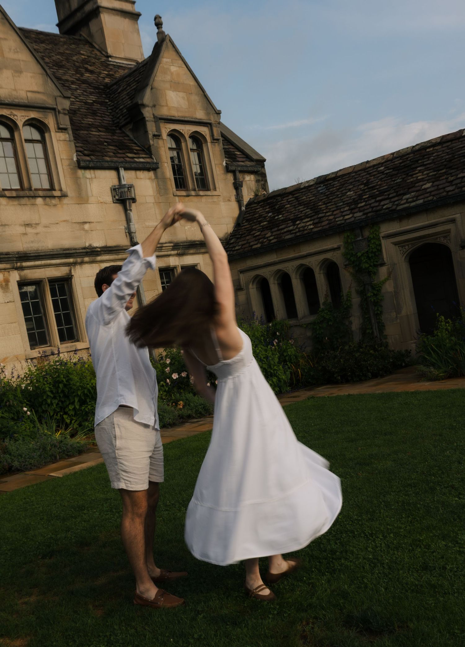 Engaged couple twirling together on grass in front of a grand stone English manor at dusk.