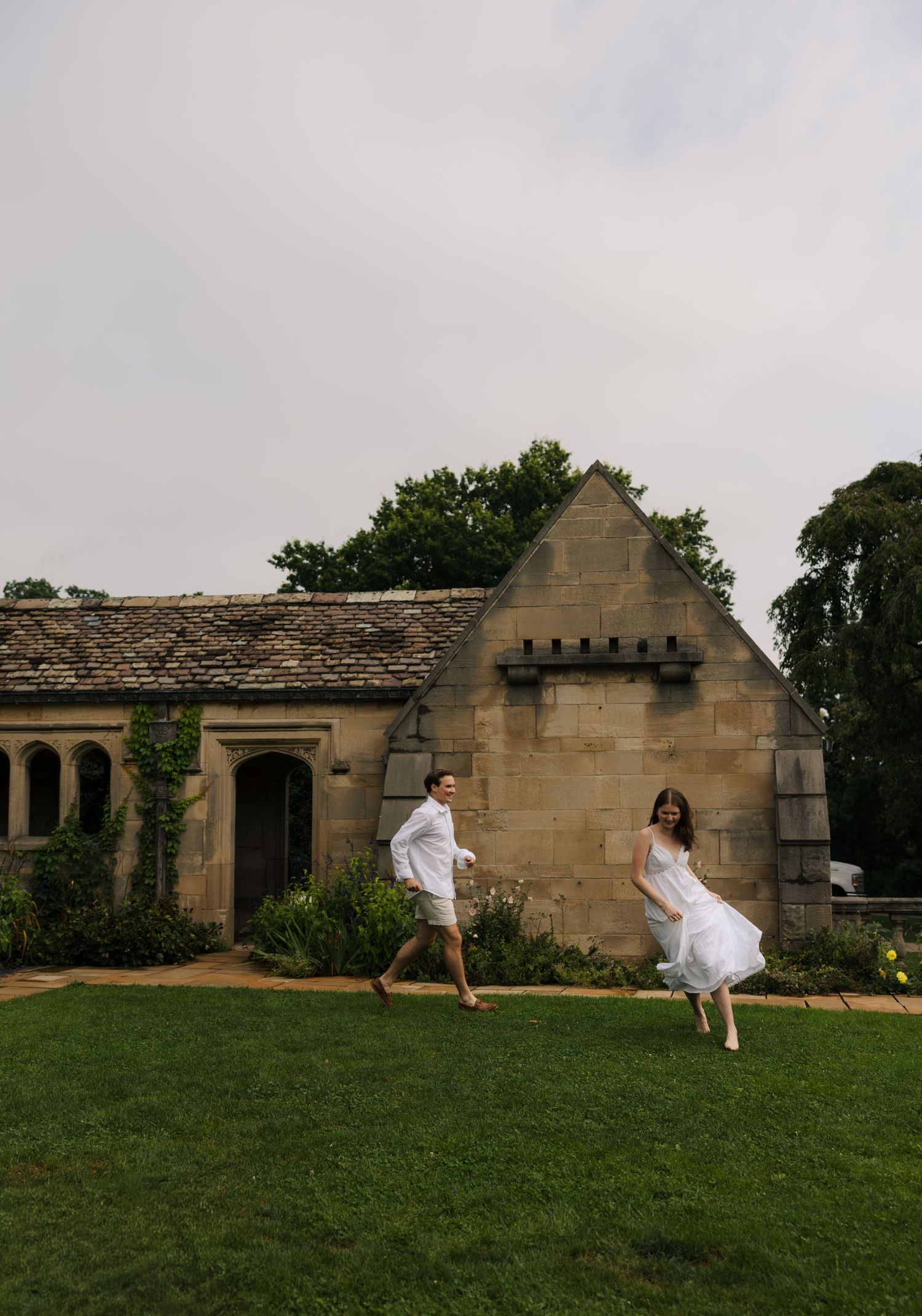 Joyful couple in white running across green lawn before stone building.