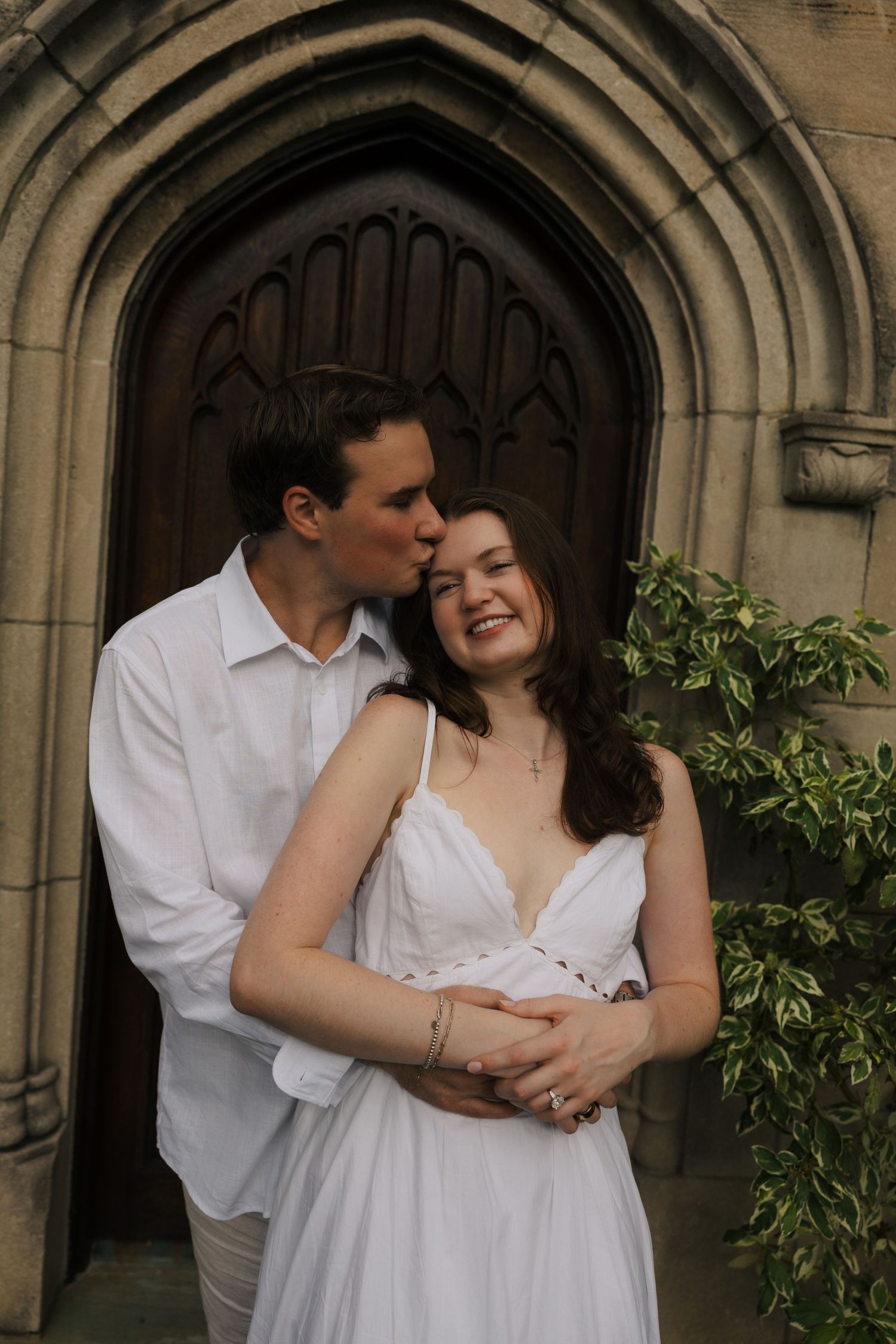 Couple in white embracing before ornate church door, man kissing womans temple as she smiles joyfully.