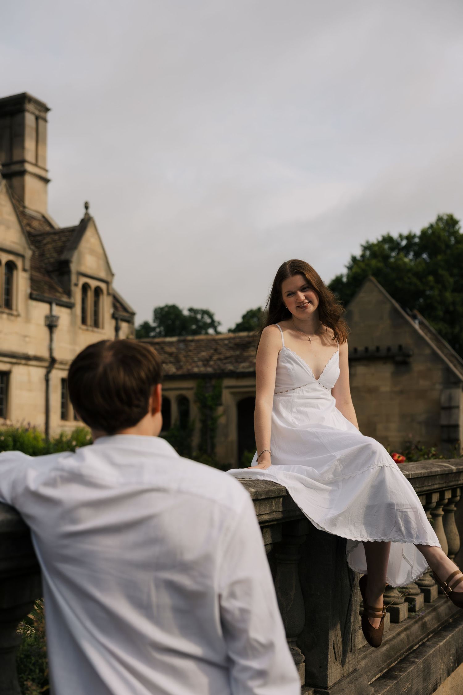 Woman in white sundress sitting on stone balcony railing smiling at man in white shirt at historic estate.