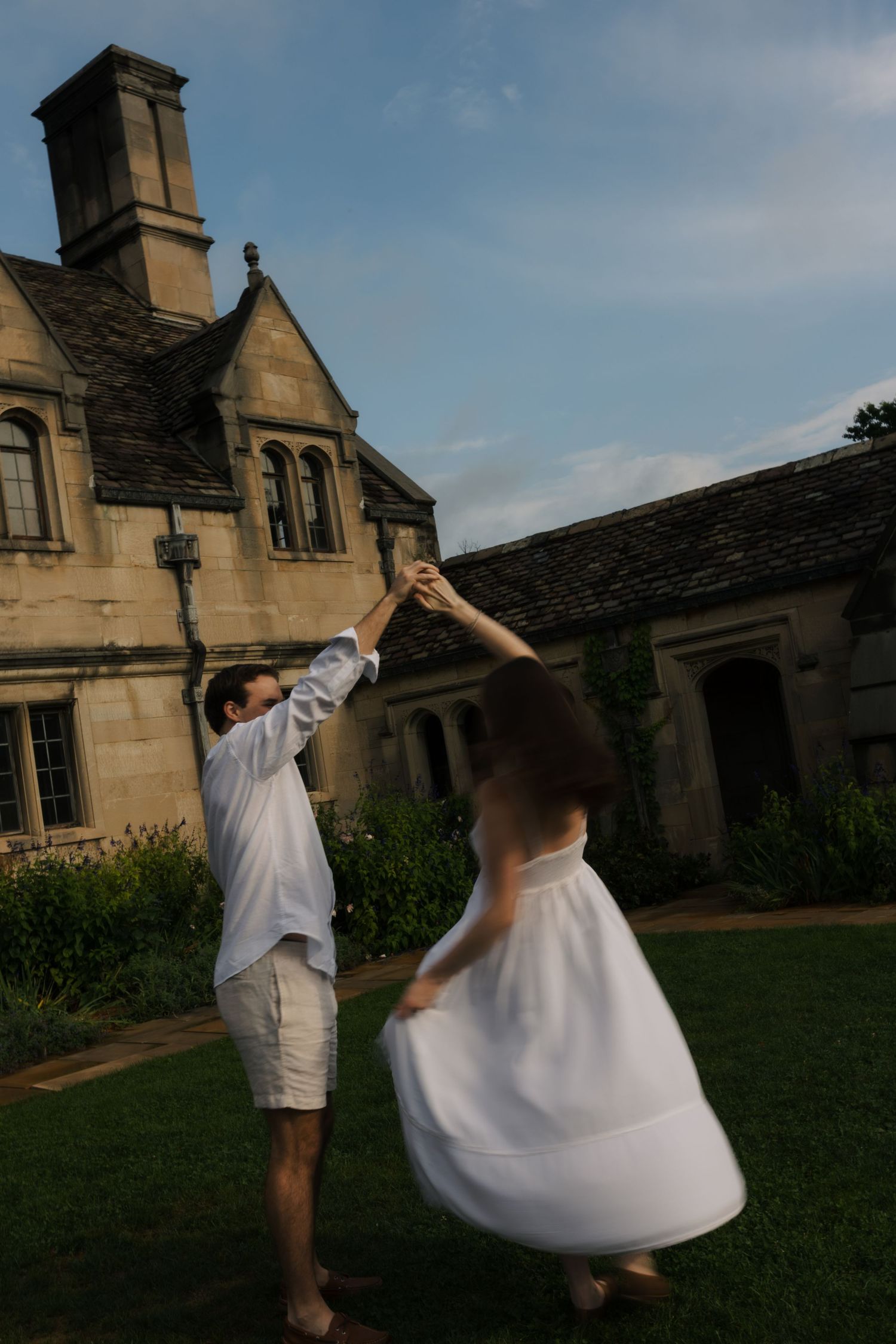 Couple spinning together outdoors in white outfits before a historic stone estate.