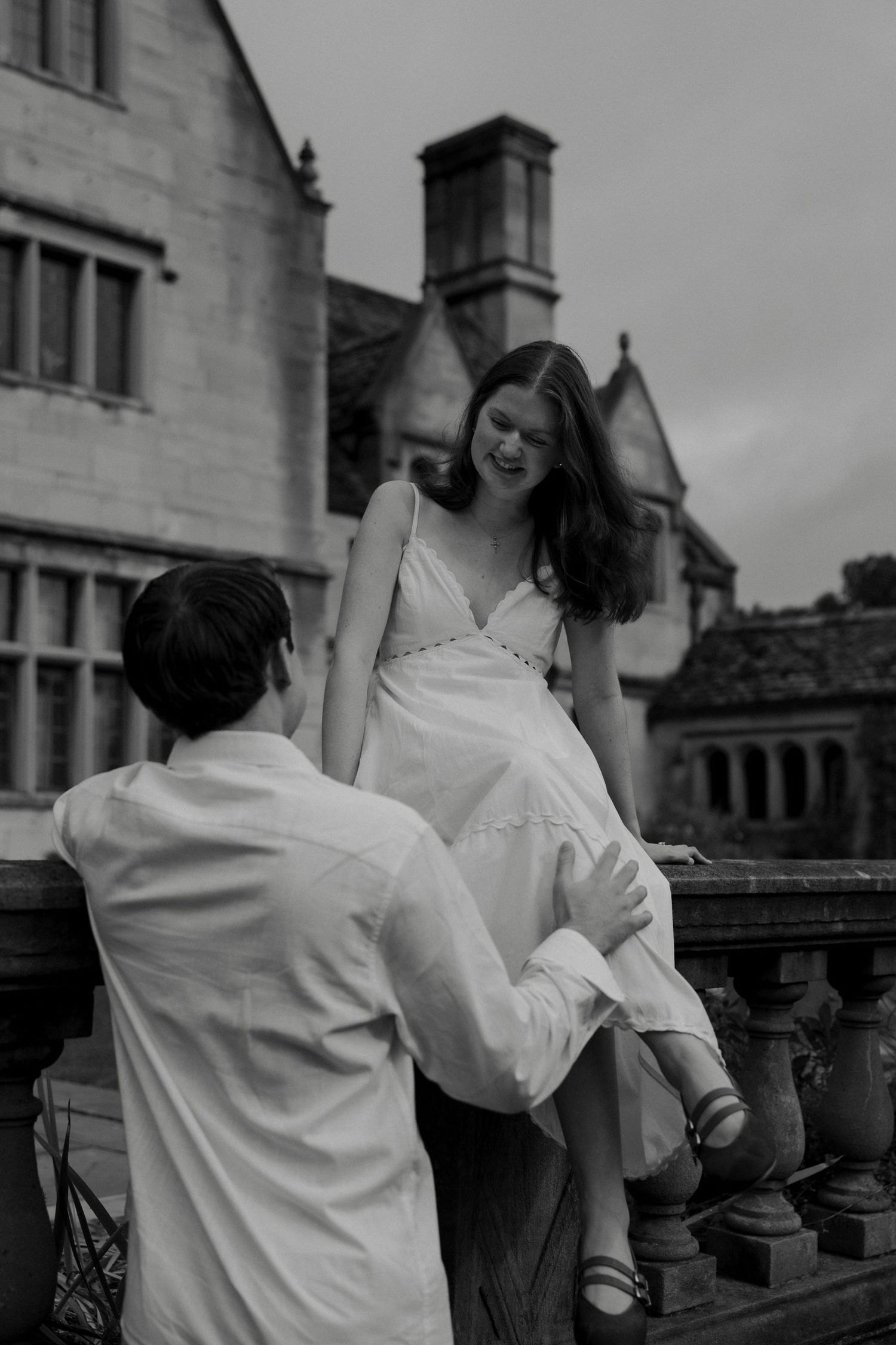 Black and white photo of a couple at a stone balustrade, woman in white dress sitting while man holds her hand.
