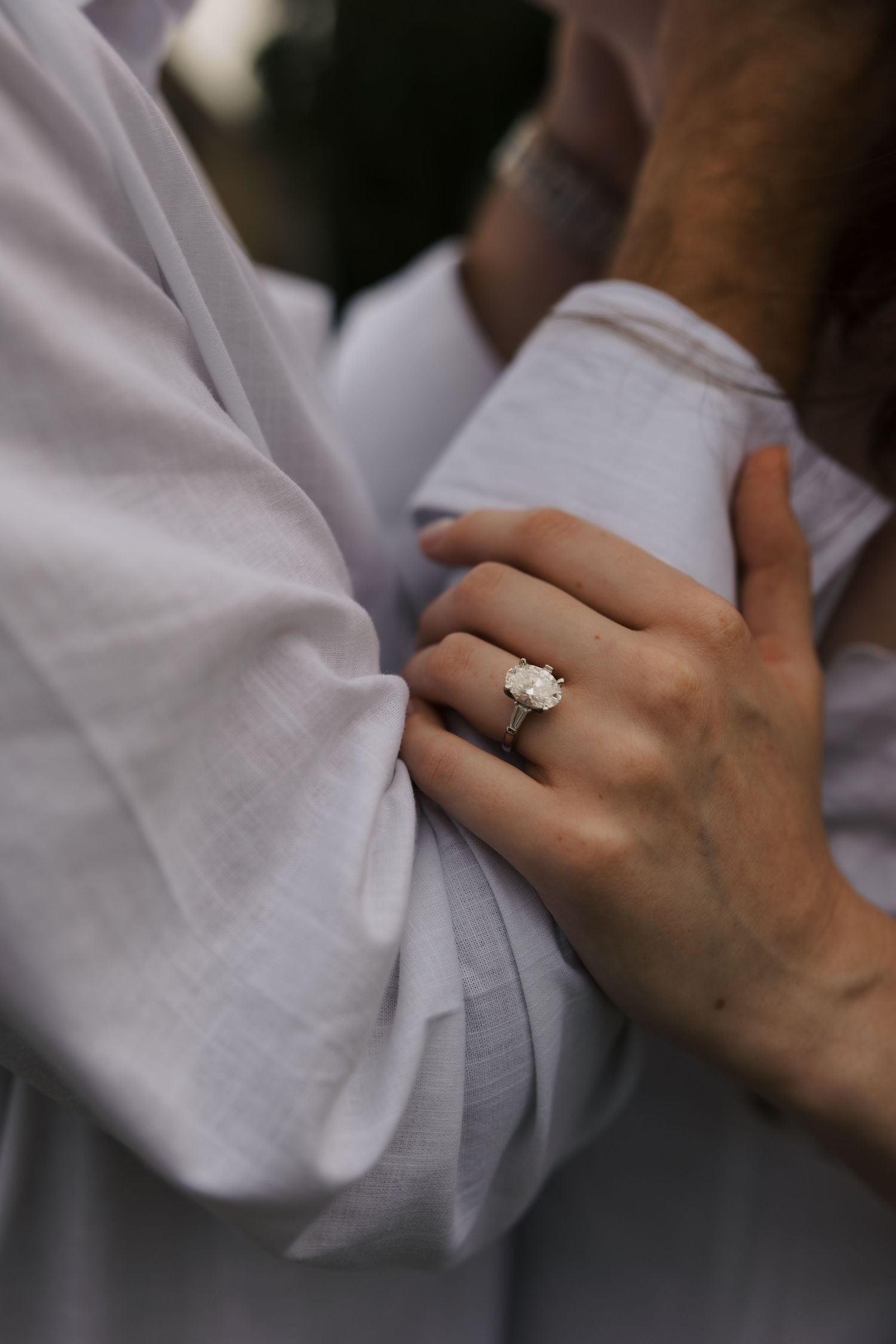 Close-up of womans hand with elegant diamond engagement ring resting on mans white linen shirt.