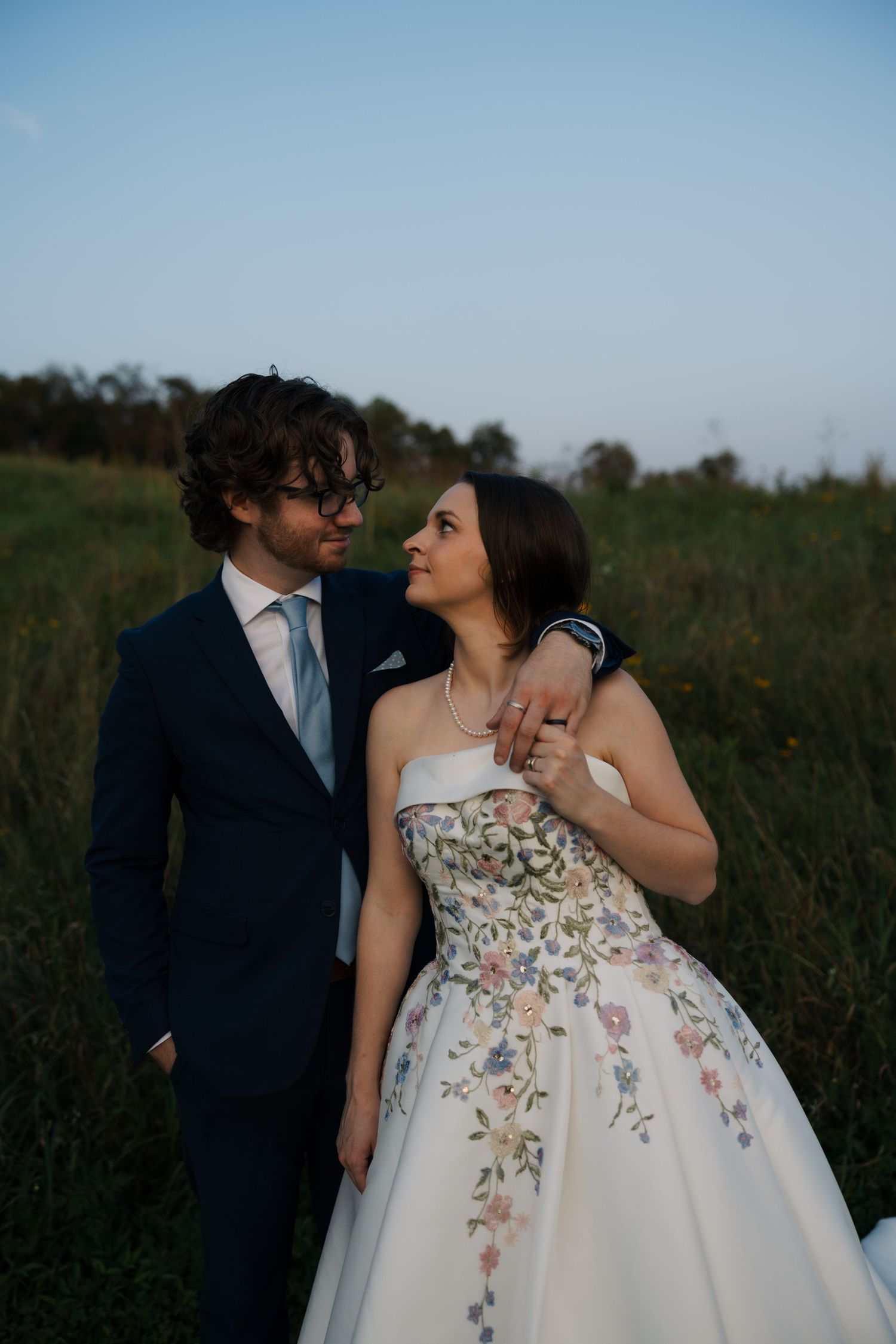 Bride and groom gazing lovingly at each other in a field at dusk, bride in a floral embroidered strapless ball gown.
