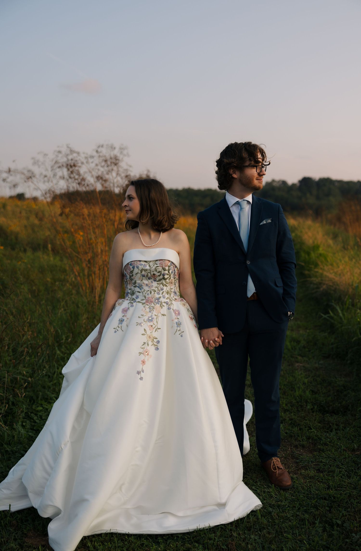Bride in floral strapless ball gown holds hands with groom in navy suit on a grassy path at golden hour.