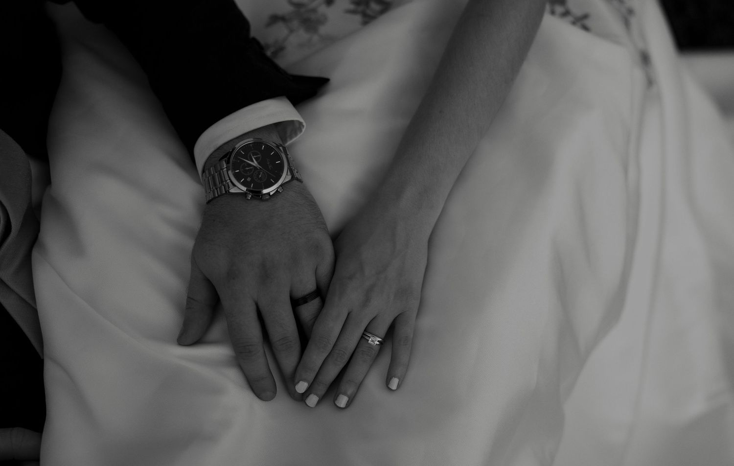Close-up black and white photo of bride and groom's hands showing wedding rings and a classic wristwatch.