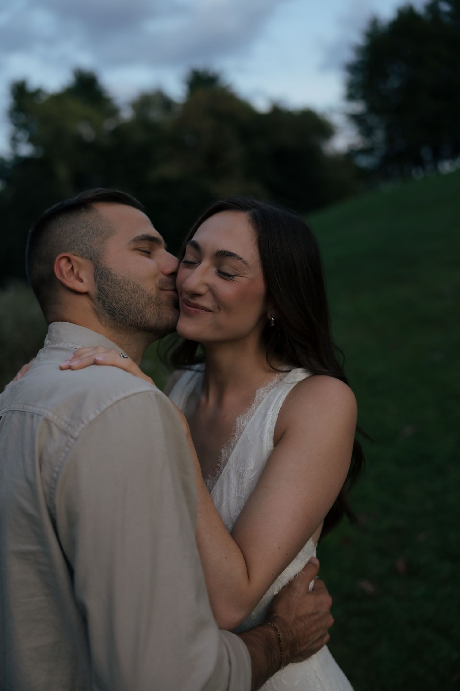 Groom kissing bride's cheek as she smiles with eyes closed in outdoor evening setting.