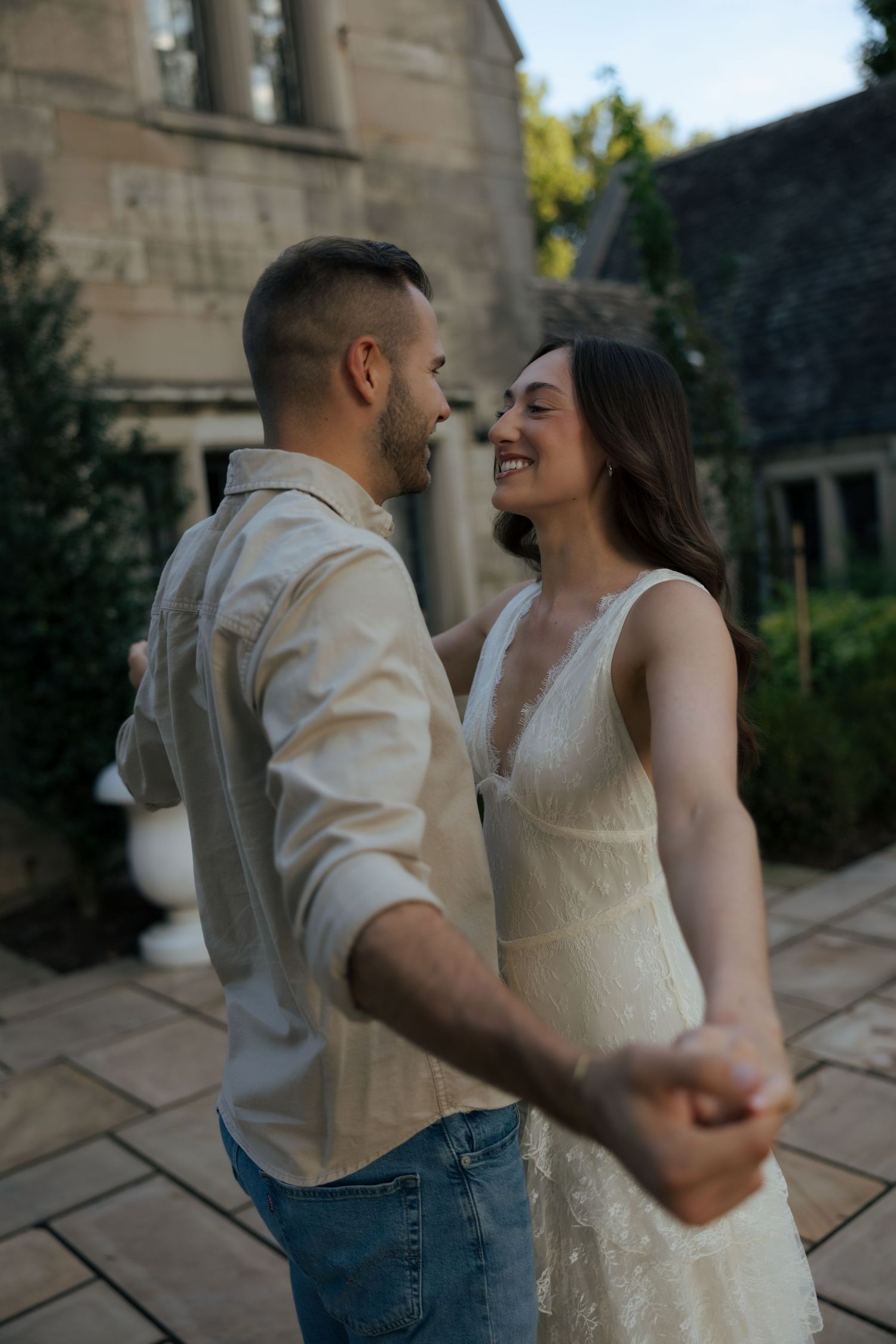 A smiling couple dances together outdoors, the woman in a white lace dress and man in a beige shirt and jeans.