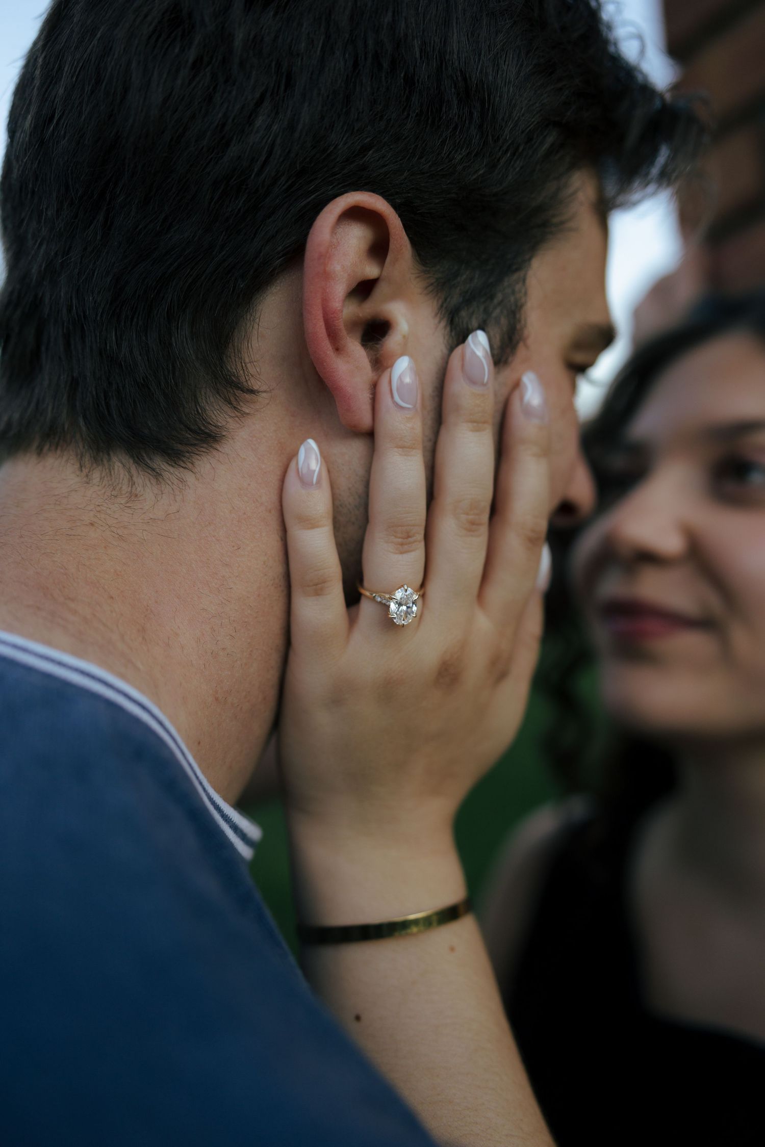 Woman wearing diamond engagement ring tenderly cupping man's face, smiling joyfully in romantic outdoor setting.