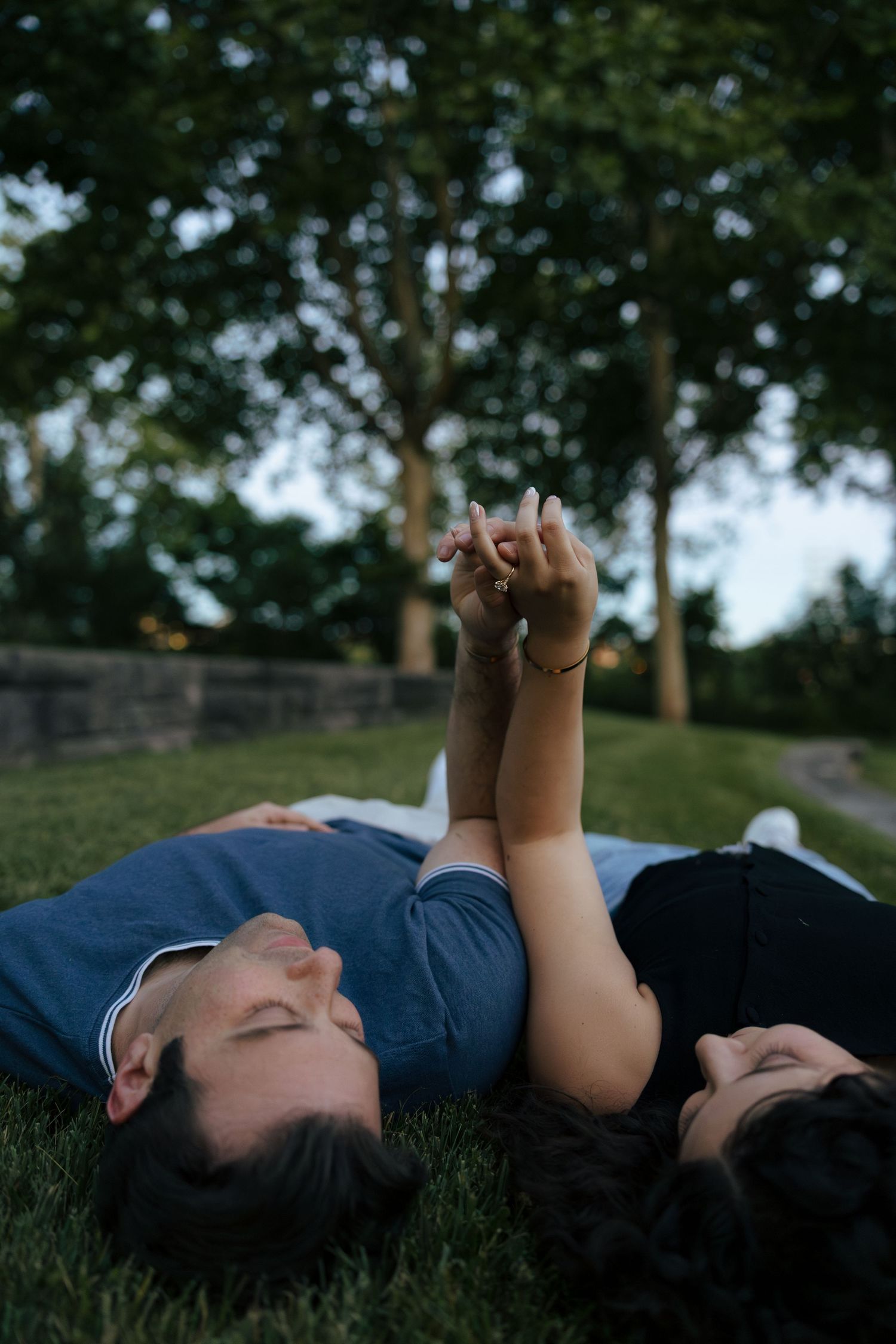 Couple lying on grass holding hands raised toward sky in romantic park setting.
