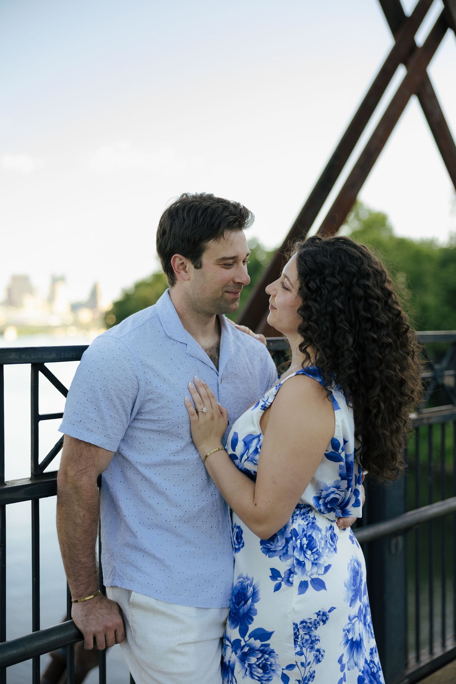 Couple gazing lovingly at each other on a bridge with city skyline in background.