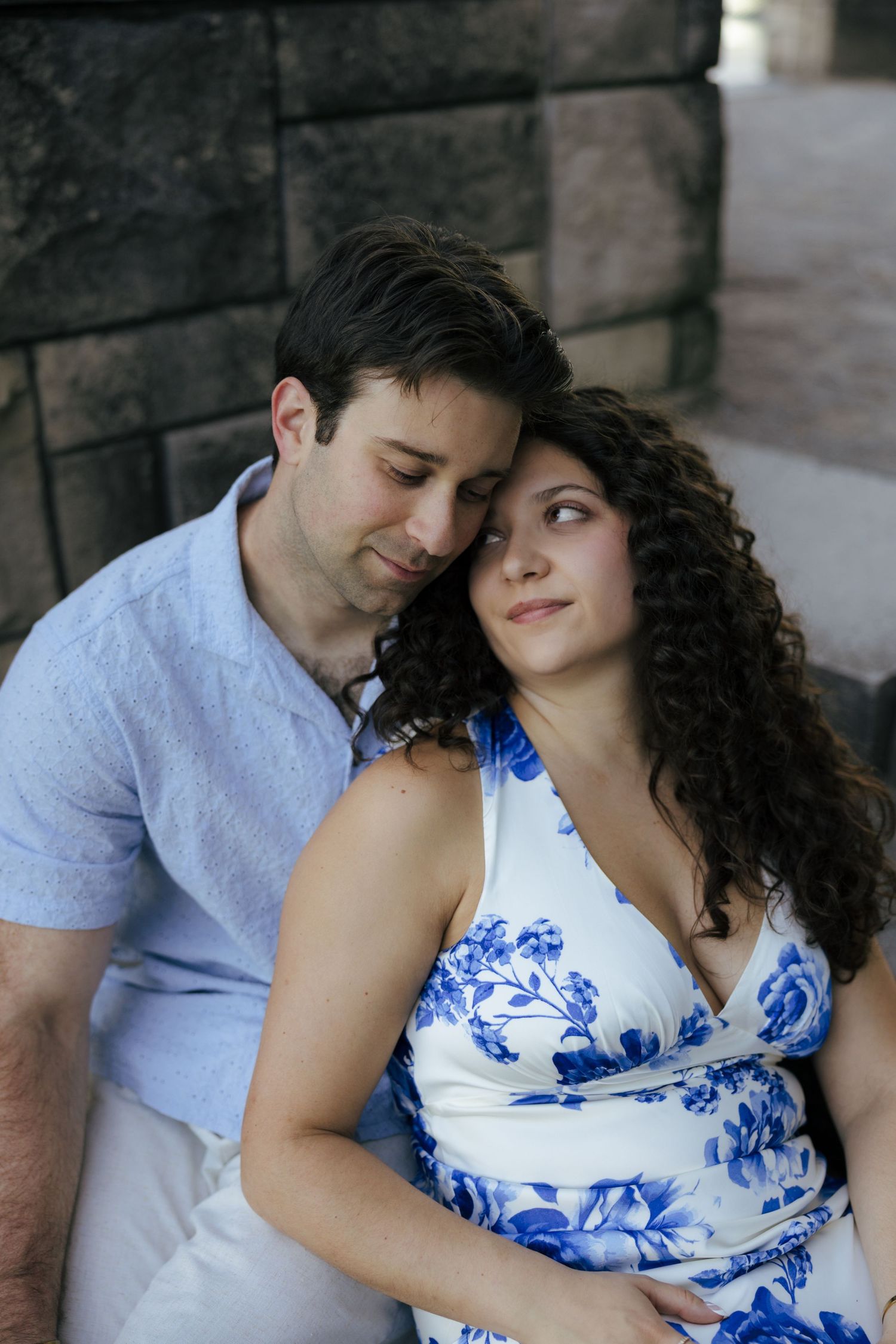 Romantic couple sitting together, woman in blue floral dress leaning on mans shoulder against stone wall.