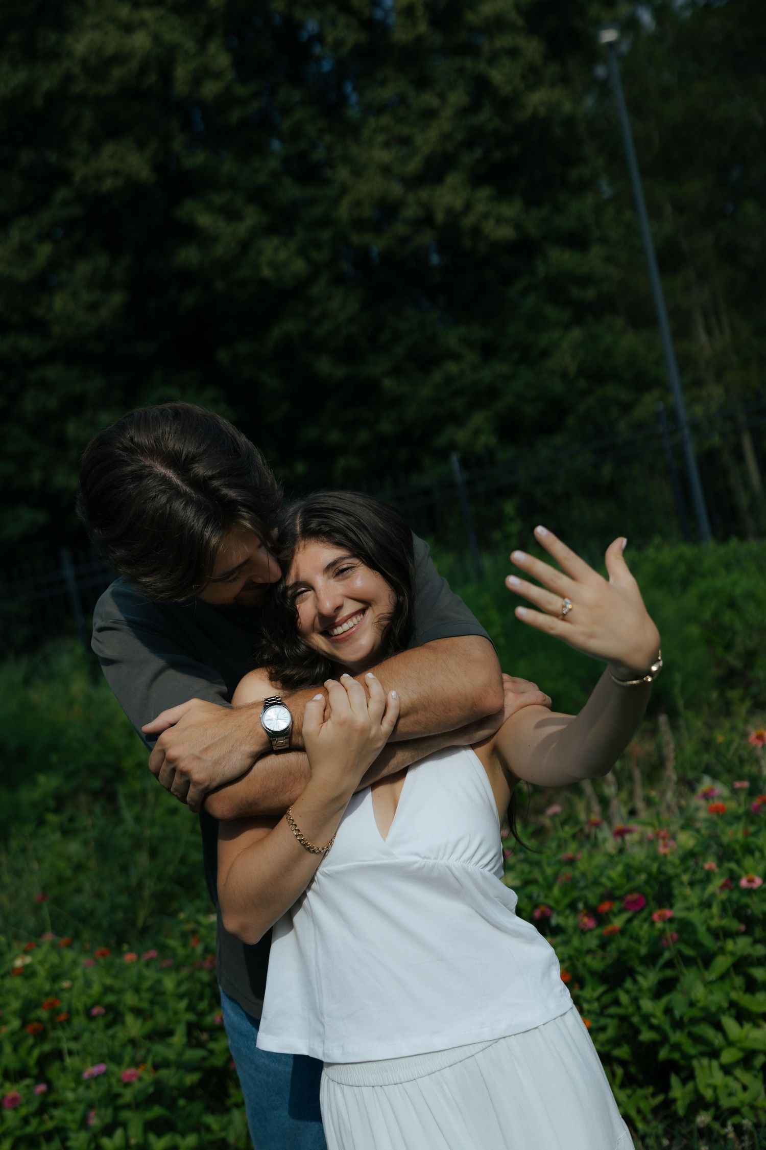 A man hugs a smiling woman from behind in a garden as she shows off her engagement ring to the camera.