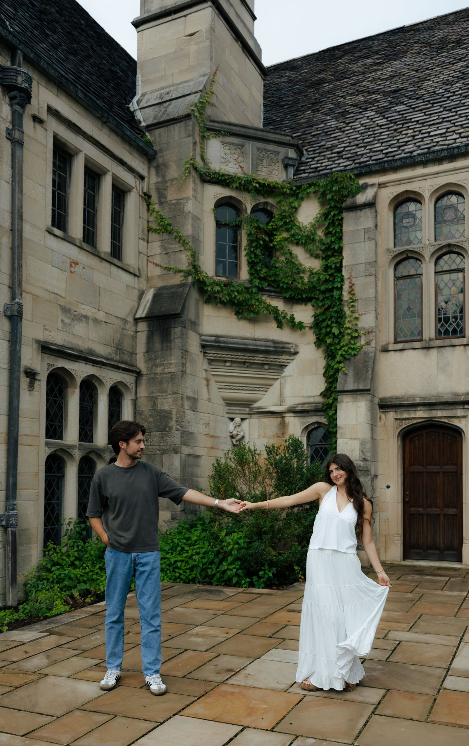 A couple holds hands at arm's length in a stone courtyard with ivy-covered building.