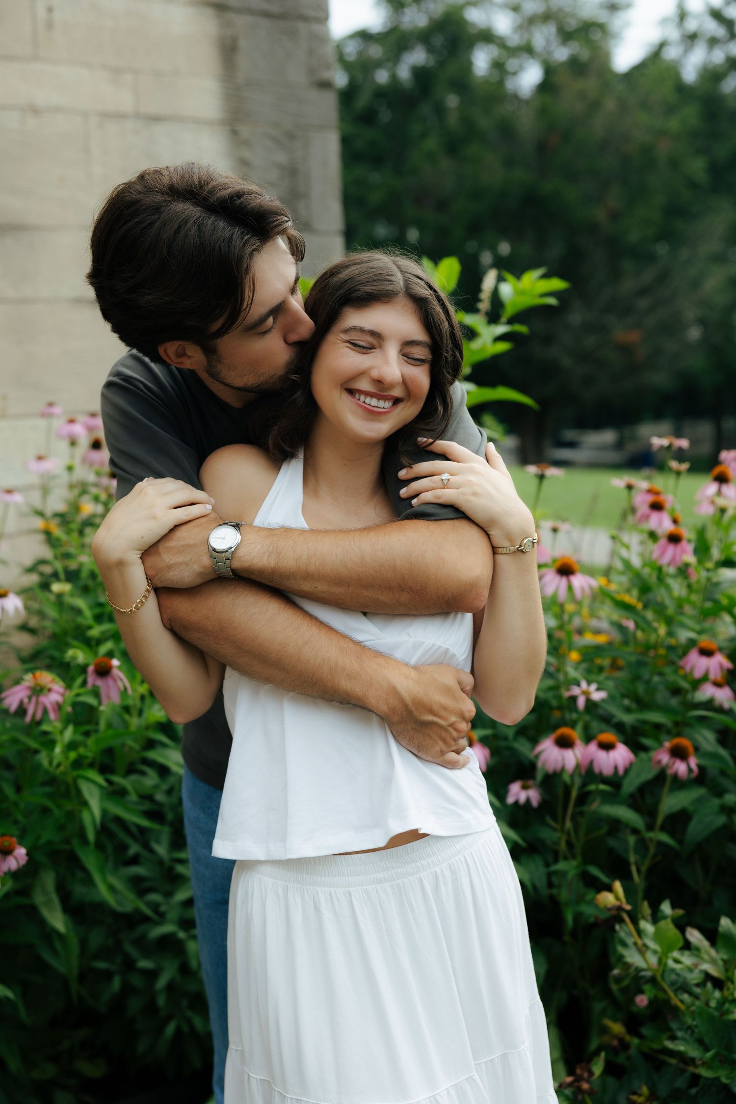 Man hugging woman from behind as she laughs joyfully in front of colorful garden flowers.